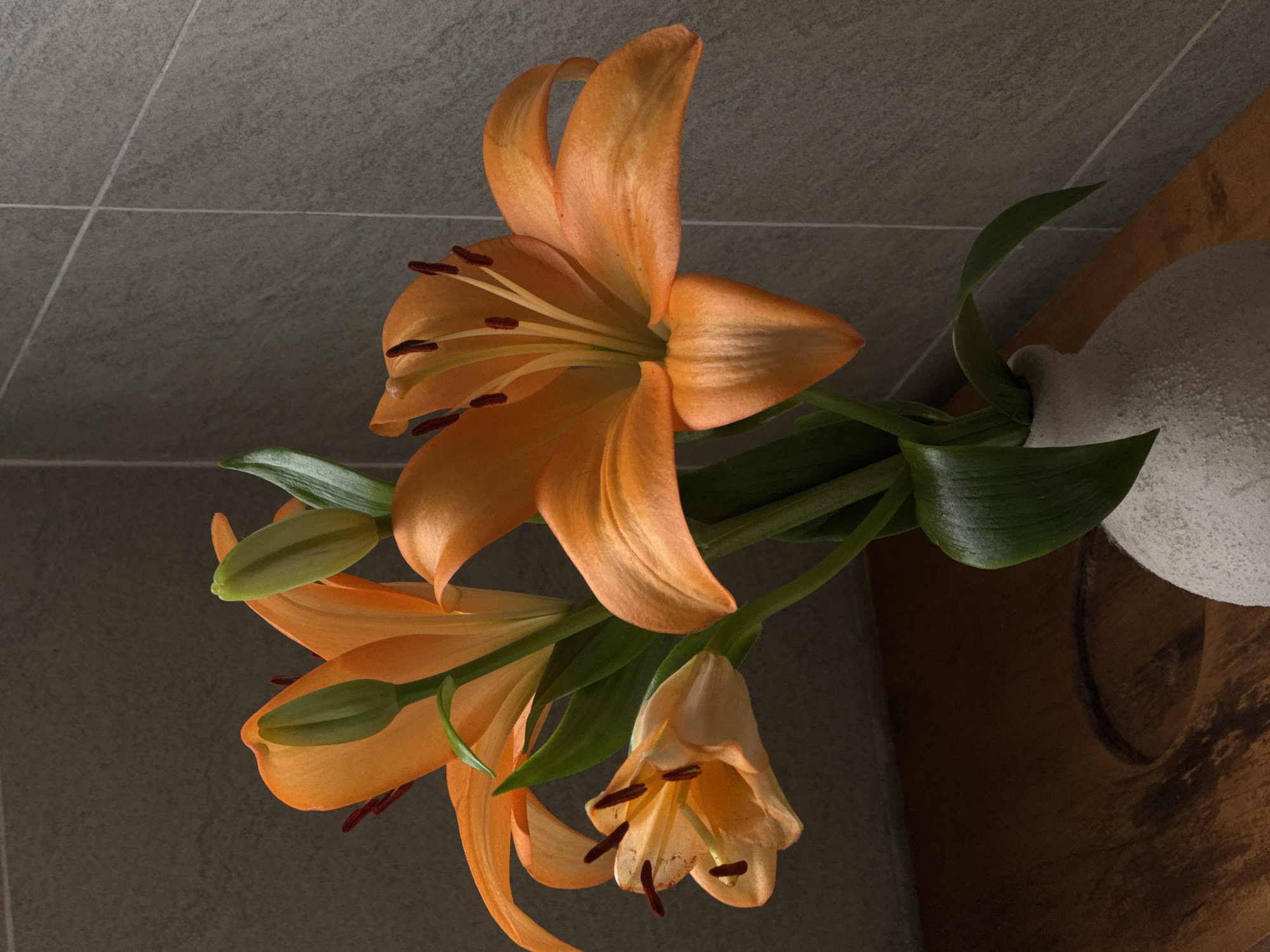 An arrangement of orange lilies in a textured white vase sits on a wooden surface against a grey tiled wall. The lilies, with their distinct petals and dark stamen, are the central focus. Two lilies are in full bloom, while a third is partially open and two green buds are visible. The wooden surface appears to be a tabletop or shelf, with a natural, possibly carved, texture. The background wall is composed of large, rectangular grey tiles with visible grout lines. The lighting suggests an indoor setting, possibly a studio or a home, with soft shadows cast by the flowers. There are no people present in the image.
