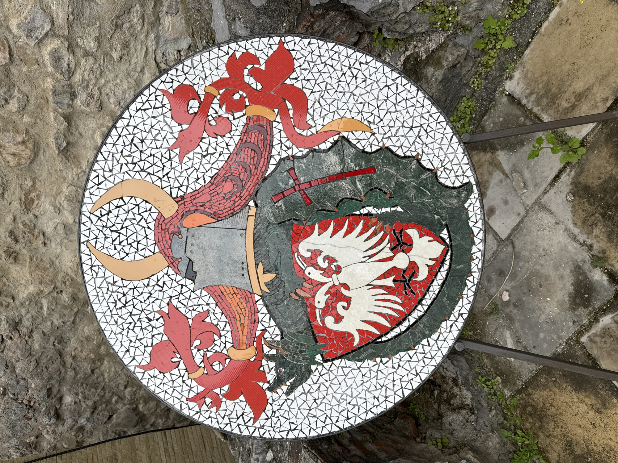 The image depicts a mosaic table top outdoors in Despotovac Municipality, Serbia. The mosaic features a coat of arms with a helm, dragon, and shield containing white birds on a red background. The background of the mosaic is white with triangular tesserae. The table is set against a backdrop of stone walls and paved surfaces, with small plants growing nearby.