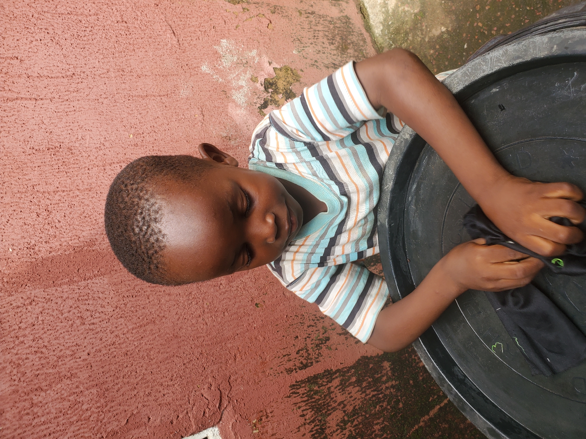 A young boy is leaning into a large black tub, with his head bowed and eyes closed, appearing to be concentrating on something inside the tub. He is wearing a striped t-shirt with horizontal bands of white, light blue, orange, and black. His hands are inside the tub, holding onto a piece of black fabric. The tub is set against a textured reddish-brown wall, with some patches of green moss or algae visible on the ground around it. The scene suggests an outdoor setting, possibly in a residential area, given the wall and the tub. The lighting indicates it is daytime. The overall mood seems to be one of quiet focus or perhaps a moment of rest for the child. There is no visible text in the image.