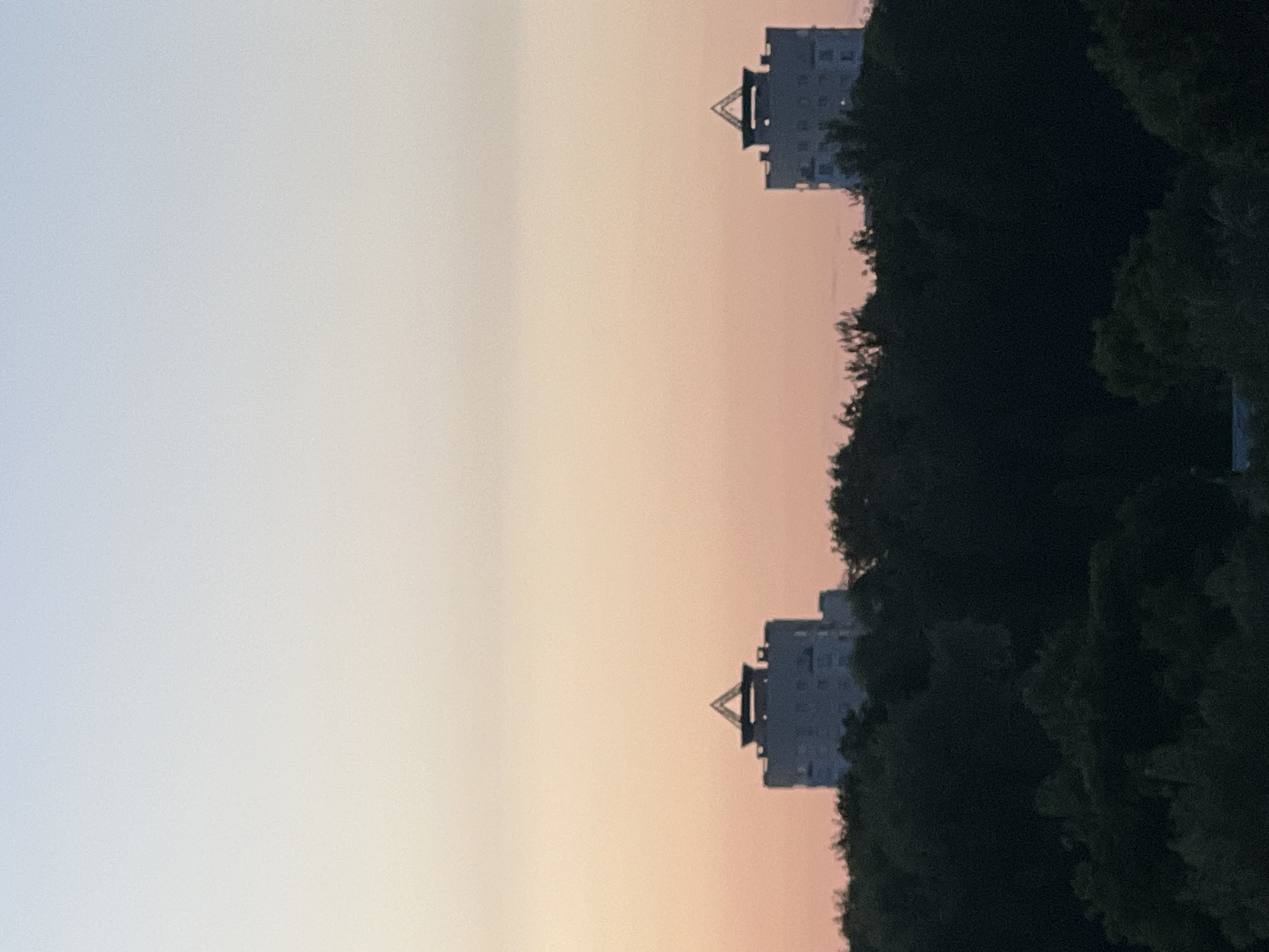The image captures a tranquil twilight scene over a city, possibly Minsk, Belarus, as indicated by the presence of identical, distinctive apartment buildings. The sky transitions from a pale blue at the top to a soft peachy-pink horizon. Silhouetted against this colorful sky are two tall, multi-story apartment buildings, each topped with a unique, angular roof structure resembling an inverted pyramid. These buildings are positioned symmetrically in the mid-ground, separated by a significant distance.
In the foreground and between the buildings, a dense, dark mass of trees forms a natural barrier, obscuring the lower portions of the buildings and the ground below. The trees are rendered in deep shadows, their foliage appearing almost black, suggesting the deepening dusk. The overall impression is one of urban development harmonizing with a natural landscape, framed by a serene, softly colored sky. There are no people or immediate actions depicted; the scene is static and peaceful.