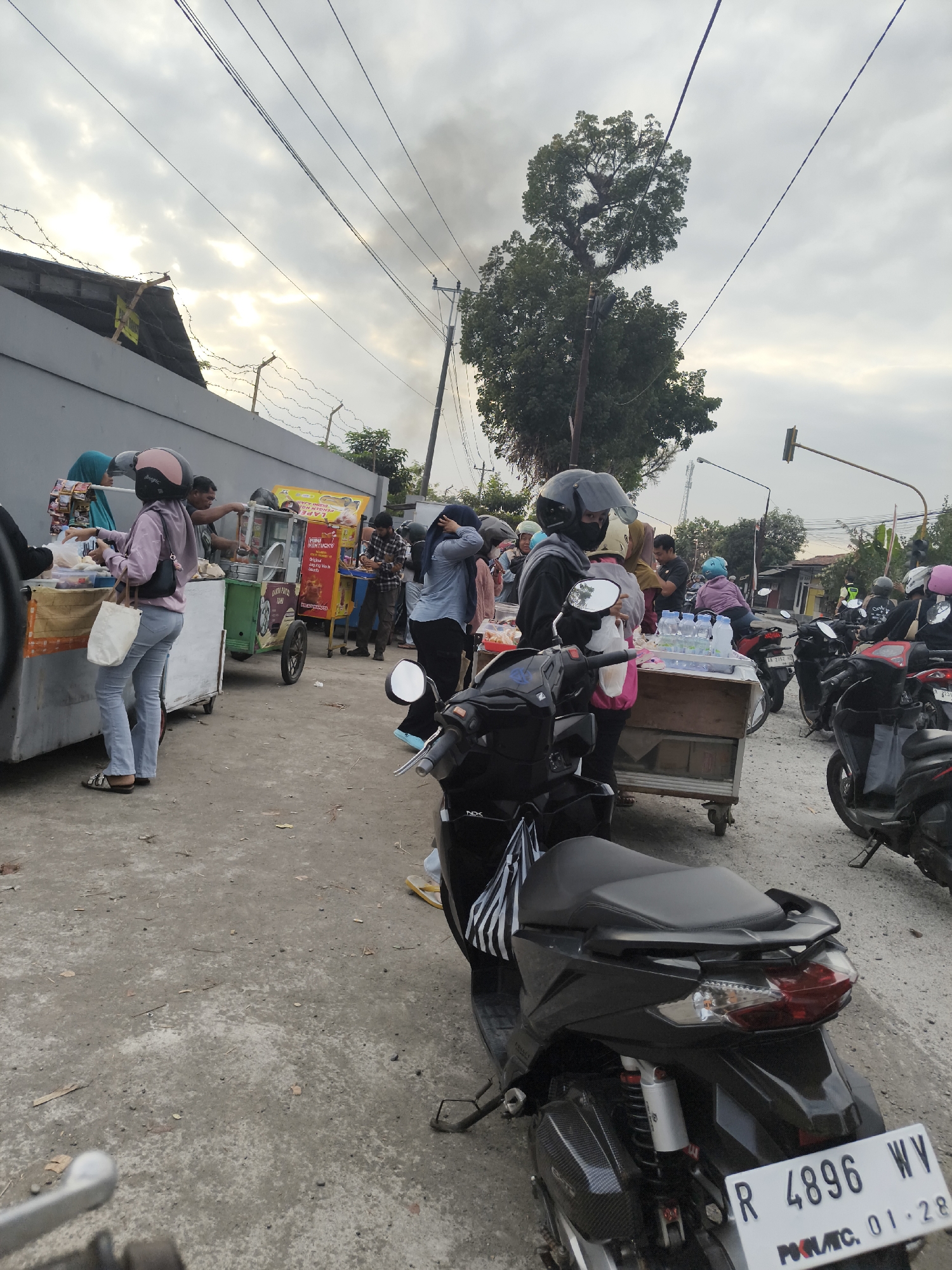 A street food vending scene is depicted in Bukateja, Indonesia, with a focus on motorcycles and food carts. In the foreground, a black motorcycle with a license plate reading "R 4896 WV" is parked. Behind it, a woman wearing a hijab and a pink helmet is interacting with a food vendor at a cart. Several other people are visible, some on motorcycles and others at food stalls. The environment appears to be an outdoor market or roadside setting, with a concrete ground and a cloudy sky overhead. The overall atmosphere suggests a typical day at a bustling local marketplace.