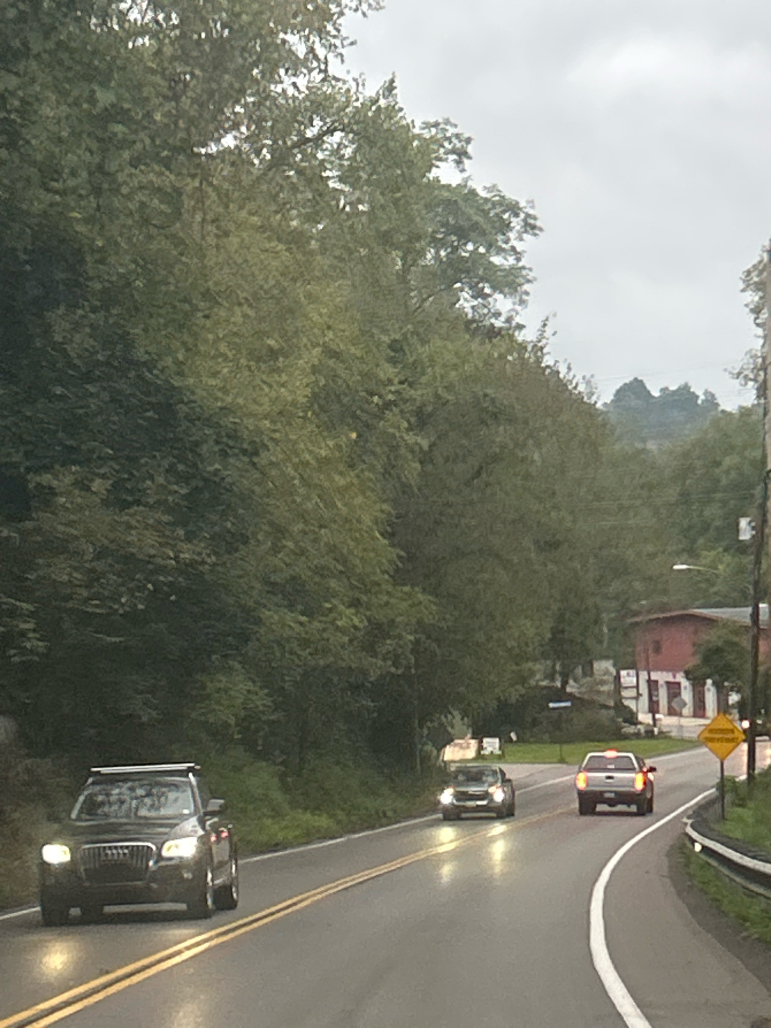 The image depicts a view from a vehicle traveling on a wet asphalt road in Scott Township, United States of America. The road has double yellow lines indicating a two-way street. Three vehicles are visible on the road. In the foreground, a dark-colored SUV with its headlights on is driving in the left lane. Further ahead, a dark sedan is in the right lane, also with headlights on, and a white pickup truck is in front of it, also with its brake lights illuminated. The environment is characterized by dense green trees on the left side of the road and a sloped hillside on the right, also covered in trees. A red building with white trim and a yellow diamond-shaped sign are visible on the right side of the road. The sky is overcast and appears grayish. There are no people clearly visible.