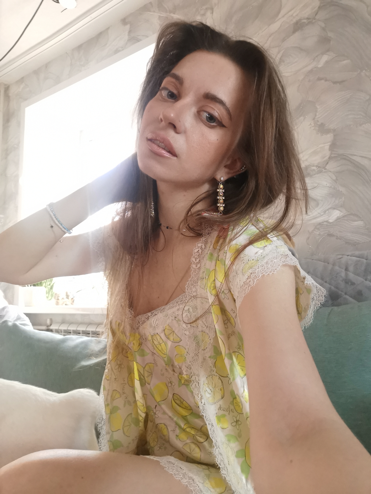 A young woman is depicted in a close-up selfie, captured indoors during bright daylight. She appears to be in her 20s or 30s, with long, wavy brown hair that frames her face. Her makeup includes winged eyeliner and glossy lips. She gazes directly at the camera with a subtle, serene expression.
She is wearing a short-sleeved top, possibly made of satin or silk, featuring a playful yellow and green lemon pattern with delicate white lace trim along the neckline and sleeves. Her jewelry includes long, elaborate dangling earrings adorned with multiple colorful stones or beads, a small stud earring, a thin choker-style necklace, and two beaded bracelets (one white, one blue and white) on her right wrist. Her right arm is raised, with her hand resting behind her head or neck in a posed manner.
The background reveals an interior setting, likely a home. A large, brightly lit window on the left side of the frame suggests daytime. Below the window, a radiator is visible. The wall behind her features a light grey wallpaper with a subtle, wavy, or marbled pattern. To her right, part of a teal or green sofa or cushion can be seen, and on the lower left, a fluffy white form suggests the presence of a pet. Visible text on her top, integrated into the pattern, appears to include words such as "good day," "dessert," and "Hossyn."