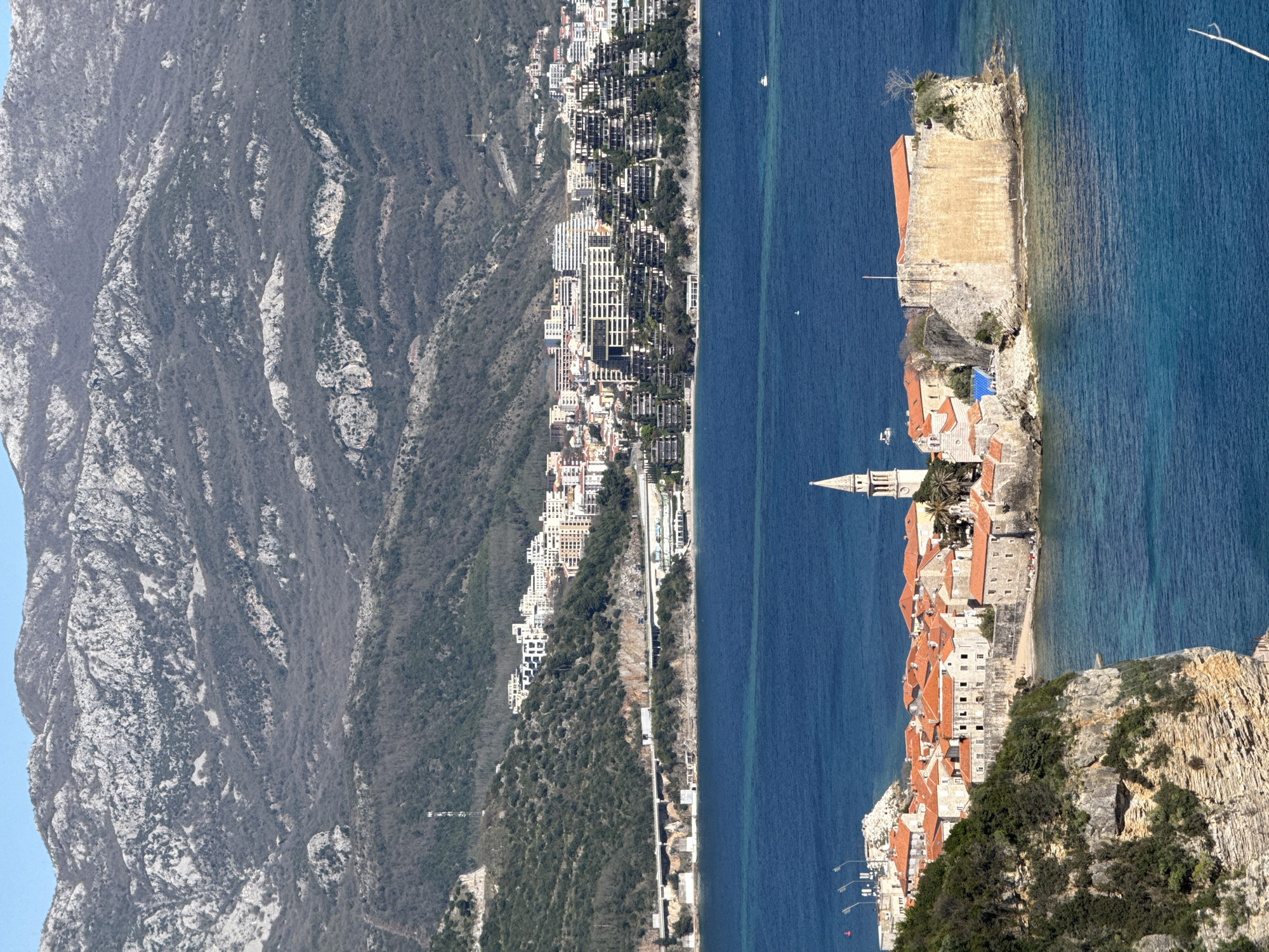 The image depicts a panoramic view of a coastal landscape in Budva, Montenegro. On the left, a mountainous terrain covered in sparse vegetation dominates the scene. Adjacent to the mountain, a line of buildings stretches along the coast, indicating a densely populated area. The color palette on this side is muted, with grays and browns dominating.
The Adriatic Sea separates the mainland from the island of Sveti Stefan, which features a cluster of buildings with distinctive red-tiled roofs and a tall church spire. The island's architecture is historic, with stone walls and a fort-like structure. The water is a deep blue, contrasting with the red and ochre tones of the island.
The image suggests a juxtaposition of natural and urban environments, with the rugged mountains and expansive sea bordering the built-up areas of Budva and the historic Sveti Stefan island.