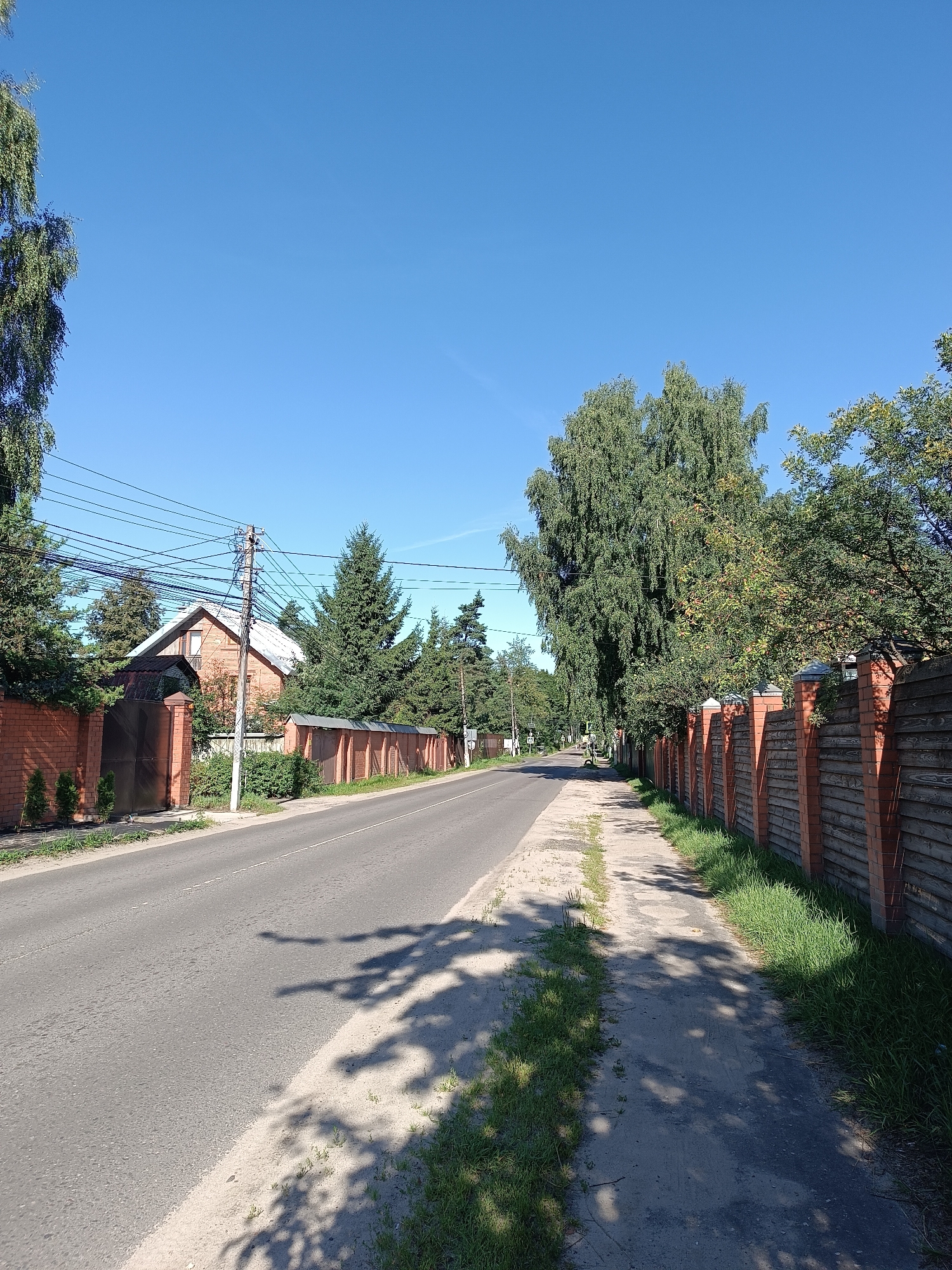 The image shows a road in Bykovo, Russia on a clear, sunny day. The road is paved and appears to be a street in a residential area, lined with fences and trees. On the left side of the road, there is a brick house with a white roof, surrounded by evergreen trees. A large brick fence with a metal gate is also visible on this side. On the right side of the road, a wooden fence with brick posts runs along the sidewalk. The sidewalk is made of dirt and grass. The sky is a clear, bright blue. Shadows of trees are cast across the road and sidewalk, indicating the sun is high in the sky. Power lines are visible strung across the scene, supported by a utility pole. There are no people or vehicles visible in the image. The overall scene depicts a quiet, suburban street in a temperate climate during daylight hours.