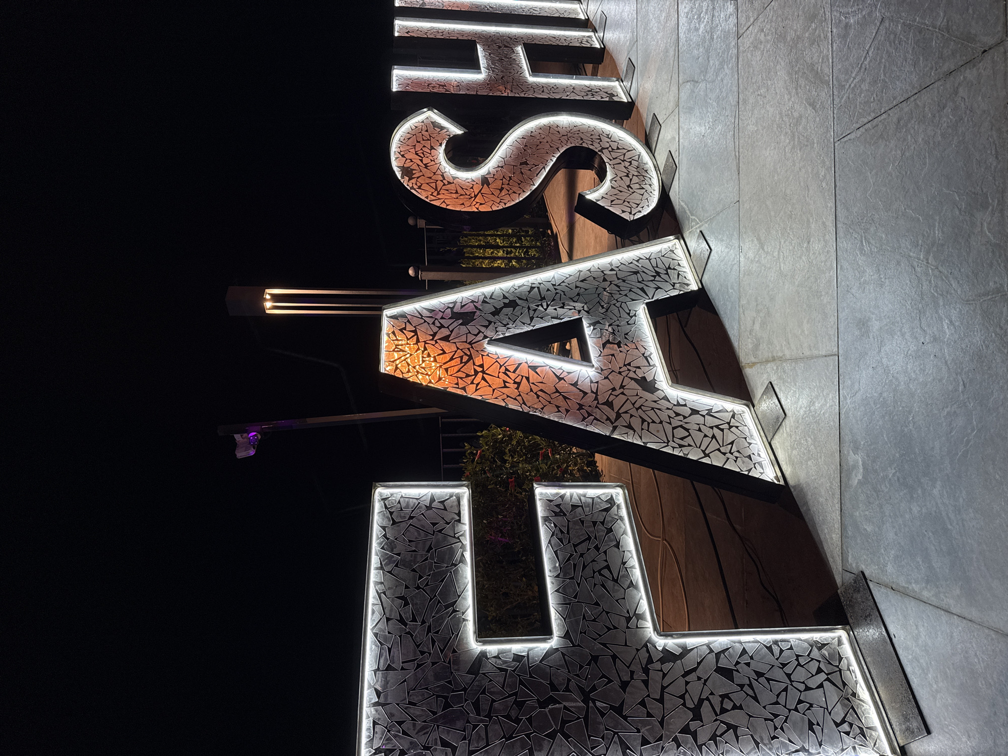 A nighttime, vertical shot depicts an illuminated sign reading "LASHS" in Kemer, Turkey. The letters are mosaic-like with internal illumination, and the camera angle is low, looking up. A security camera is visible above the "L". The sign is attached to a wall. A gray tiled surface is on the right. The background is black.