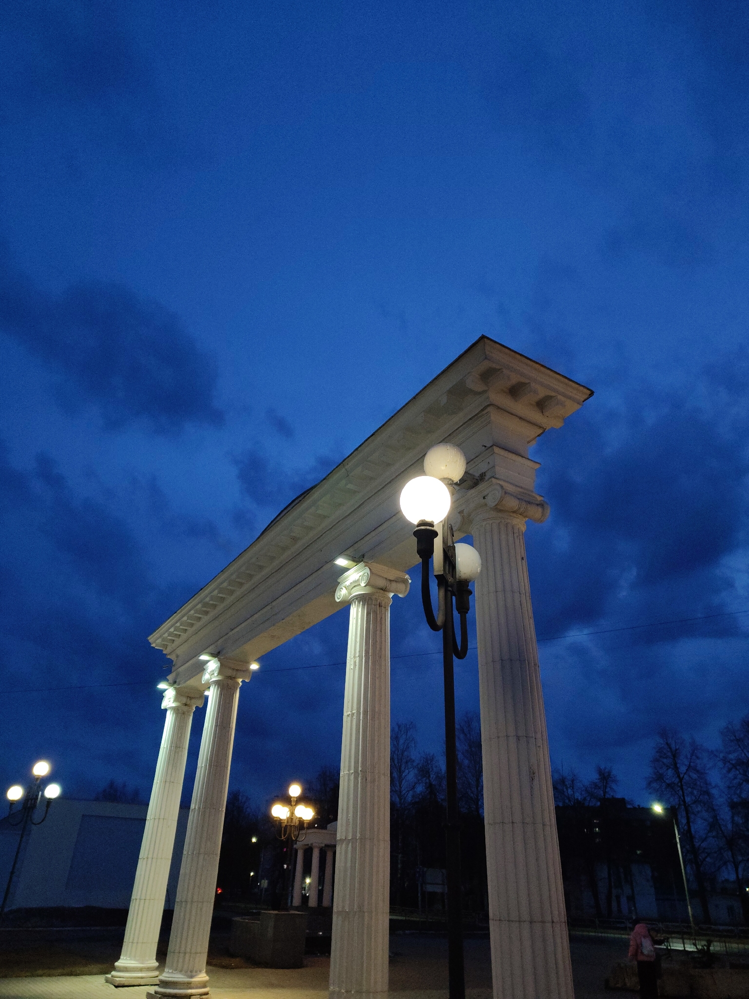 A low-angle, color photograph taken at dusk shows an archway with four white columns topped by a rectangular structure, situated outdoors in an urban setting. The archway columns have classical detailing, and the overarching structure features molded edging. To the right of the archway is a lamppost with multiple spherical lights, some of which are illuminated.
In the background, more lampposts are visible, along with trees and what appears to be a building. The sky is a deep blue, indicating the onset of night, and is marked with streaks of dark clouds.
At the lower right, a person wearing a light-colored coat can be seen. The ground appears to be paved, and the overall setting suggests a park or public square within a city. The city and state of the location are unknown.