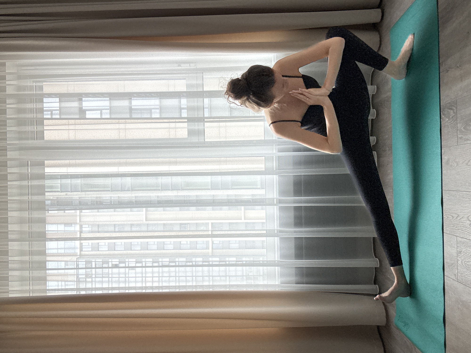 A woman is performing a side lunge yoga pose on a teal yoga mat in front of a large window with sheer white curtains and beige drapes. The window offers a view of modern high-rise buildings. The woman is wearing a black sleeveless top and black leggings. Her hands are pressed together in front of her chest in a prayer position. Her left leg is extended straight, and her right leg is bent, with her torso lowered. The floor is wooden.