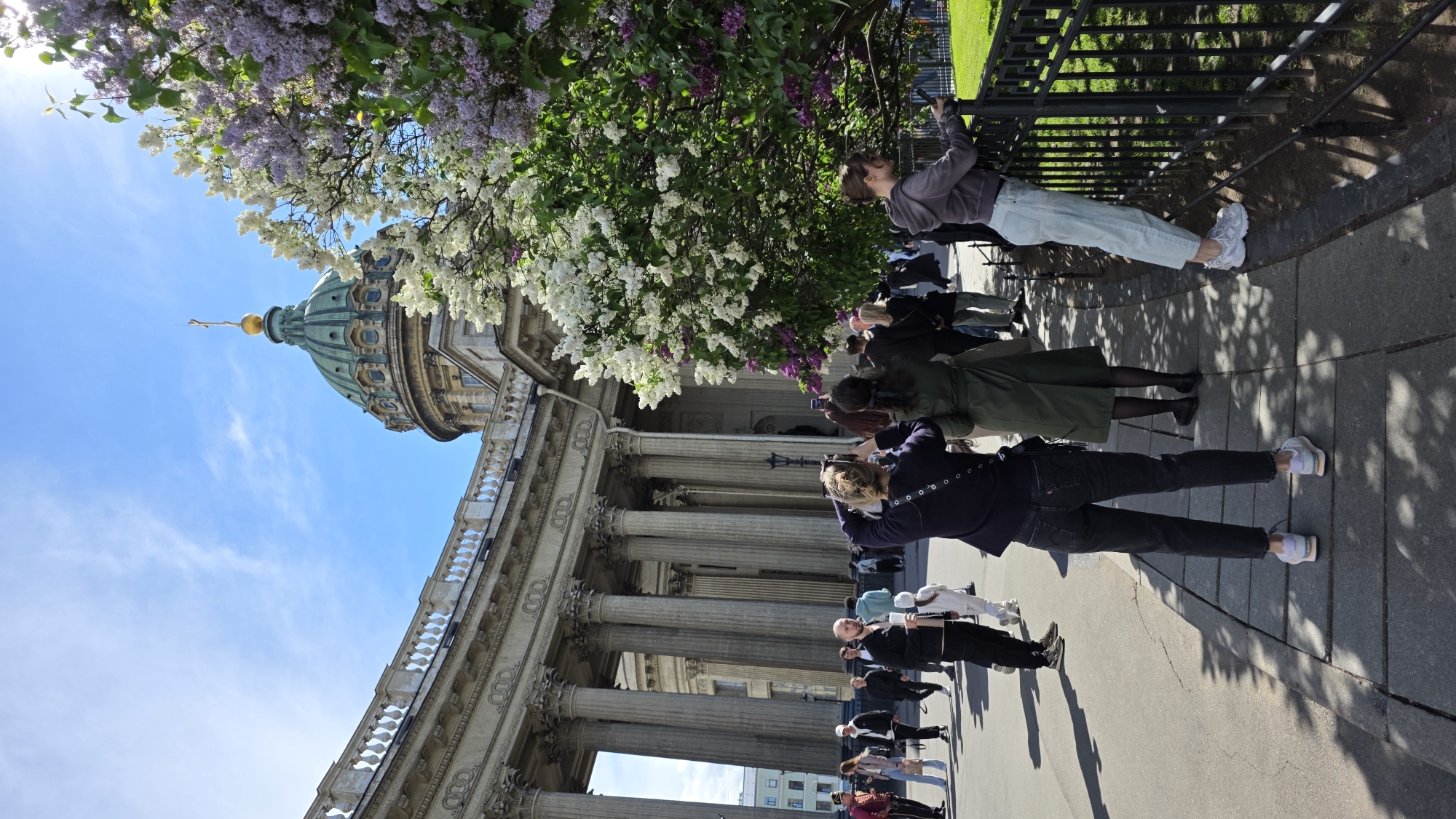 The image shows Kazan Cathedral in Saint Petersburg, Russia, from a street-level perspective. The cathedral's columns, dome, and architectural details are visible. Several people are present, some appear to be tourists taking photographs with mobile phones of the building and blooming lilac bushes. They are on a paved sidewalk next to a wrought-iron fence. The sky is blue with some clouds.