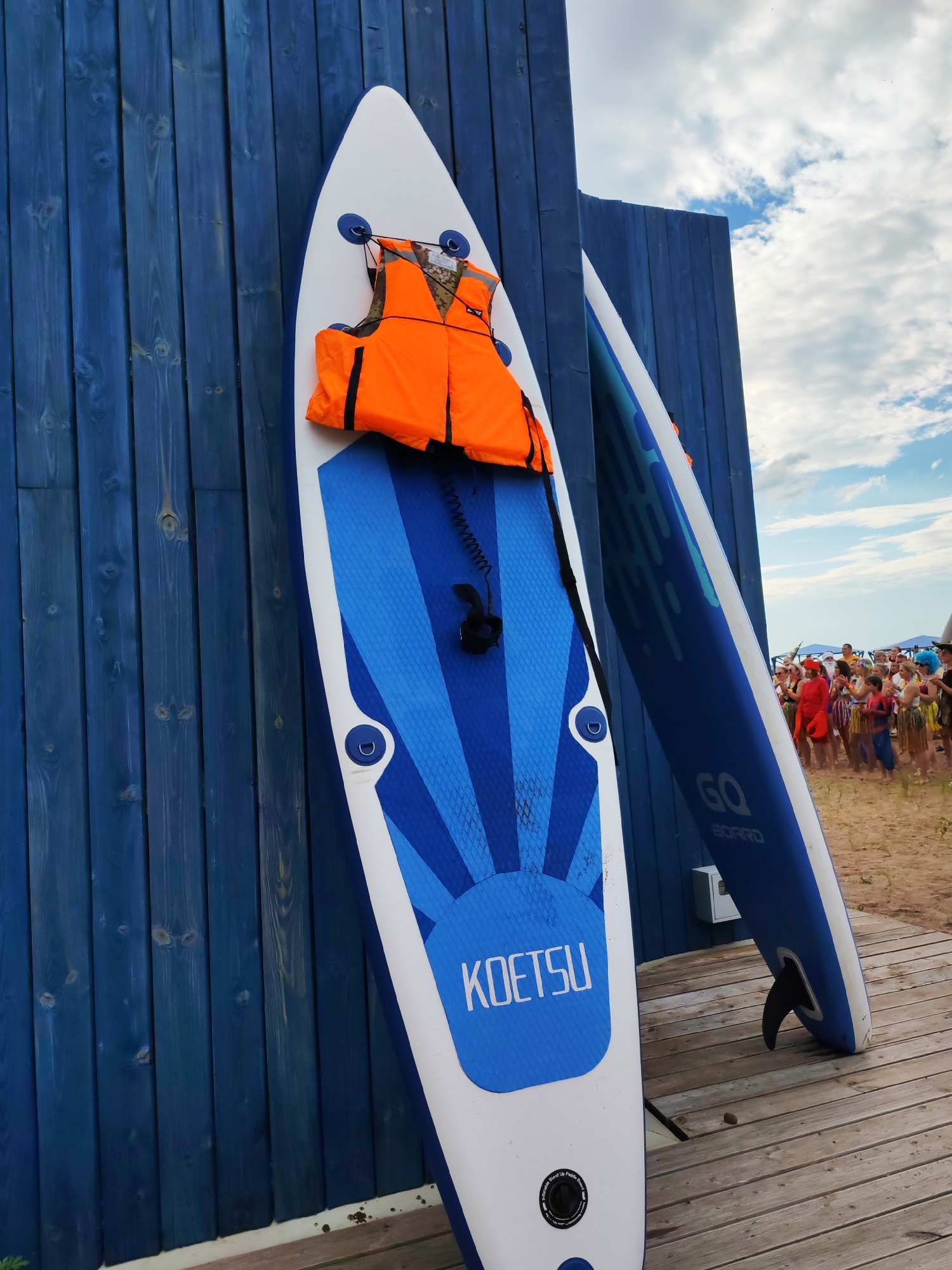 In Chaikovsky, Russia, two stand-up paddleboards are leaned against a blue wooden wall on a wooden boardwalk. An orange life jacket is strapped to the front of the closest paddleboard. In the background, a crowd of people, some dressed in costumes, gather on a sandy beach under a cloudy sky.
