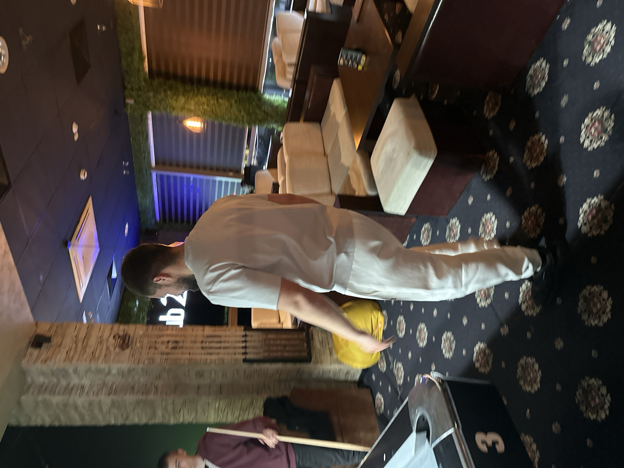 A man in a white shirt and pants is playing pool in a room with patterned carpeting, sofas, and a brick wall. Another man is holding the pool cue. The location could be a bar or recreational area in Plovdiv, Bulgaria.