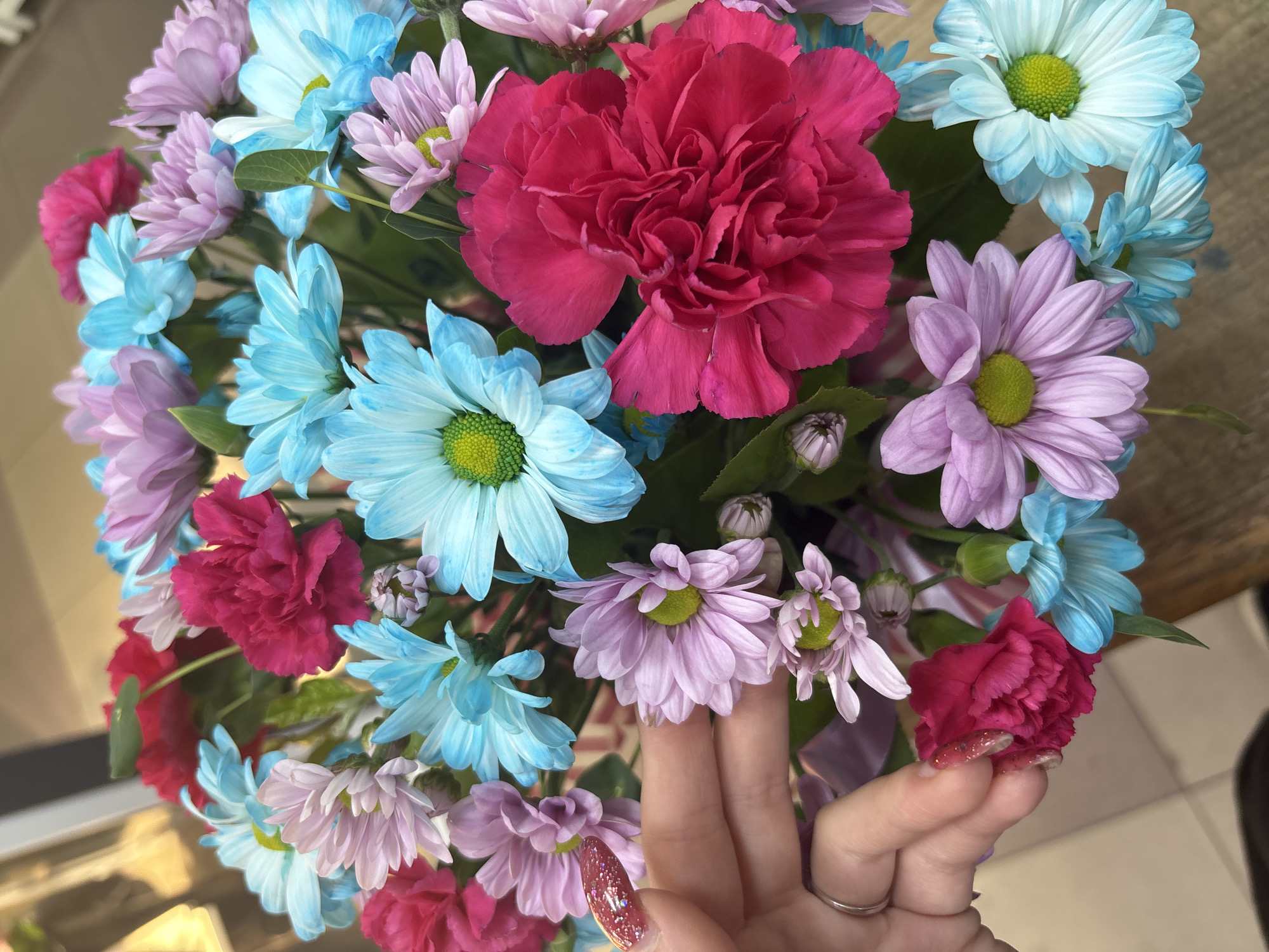 A close-up shot captures a vibrant bouquet of flowers, featuring a mix of pink and blue chrysanthemums and deep pink carnations. A human hand, adorned with a ring and sparkly nail polish, gently touches one of the pink chrysanthemums. The background suggests an indoor setting, possibly a florist shop or a home, with a blurred reflection of a window or glass display on the left and a tiled floor visible at the bottom. The lighting is bright, illuminating the colorful blooms and the delicate details of the hand and flowers.