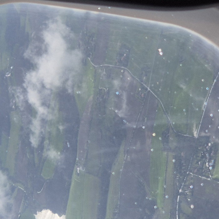 The image displays an oblique aerial perspective of a rural and semi-rural landscape located in the vicinity of Blagnac, France, observed through an aircraft window. The ground below consists primarily of variegated green agricultural fields, interspersed with areas of darker land, possibly tilled or wooded. A network of light-colored, winding roads crisscrosses the terrain. Scattered residential or agricultural structures are visible in small clusters, particularly in the lower left and central-right portions of the scene. White, diffused clouds are present, partially obscuring the ground in the upper and central sections. The aircraft window in the foreground exhibits numerous small, circular, multi-colored points of light, indicative of reflections or refractions from the window surface, alongside minor surface imperfections. No discernible individuals, specific objects beyond general landscape features, or active interactions are present within the observable scene below.