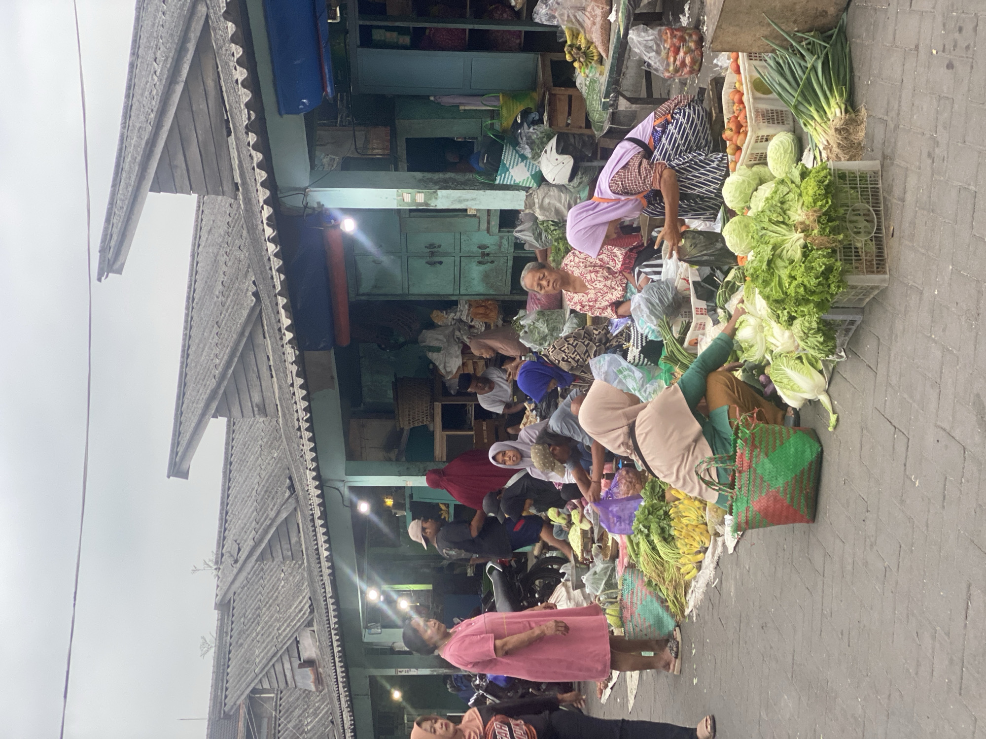 In Ambarketawang, Indonesia, an outdoor market scene unfolds under an overcast sky. Stalls with weathered, grey, corrugated roofs are arranged in a row, the architecture featuring distinctive angled wooden supports. People, mostly women dressed in traditional modest clothing and hijabs, are gathered around displays of fresh produce. Vegetables like lettuce, cabbage, onions, and bananas are piled high on tables and in woven baskets. Some women are actively selling, while others appear to be customers. The ground is paved with grey interlocking bricks. A few motorcycles are parked nearby. The overall atmosphere suggests a typical day at a local marketplace.