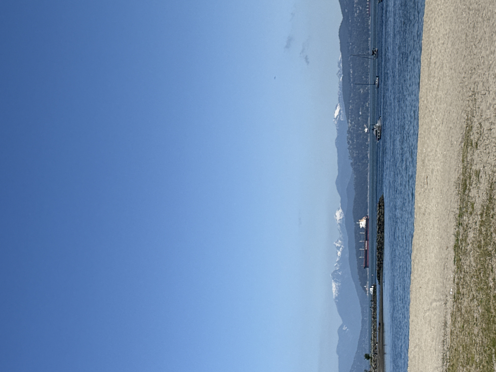 The image presents a serene panoramic landscape, oriented horizontally, depicting a sunny day in Vancouver, Canada. The top two-thirds of the frame are dominated by a vast, clear blue sky, devoid of significant clouds.
In the mid-ground, a wide expanse of deep blue water, likely part of Burrard Inlet or English Bay, stretches across the scene. Several vessels are visible on the water: closer to the foreground, a few smaller boats, possibly sailboats or pleasure crafts, are scattered. Further out, against the distant shoreline, a large cargo ship with a prominent red hull can be seen; the word "ULTRAMAR" is faintly visible on its side. A rocky breakwater or natural barrier runs parallel to the foreground, separating the immediate water from the open bay.
The horizon is defined by a majestic range of mountains, characteristic of Vancouver's North Shore. Their peaks are dusted with snow, and some atmospheric haze softens their appearance, suggesting a clear but perhaps warm day. Faint indications of urban development, such as buildings, can be seen along the lower slopes of these distant mountains.
The foreground consists of a light-colored sandy or gravelly beach, interspersed with patches of green grass or vegetation. The overall impression is one of natural beauty and urban integration. The time of day is clearly daytime, with bright, even light suggesting morning or afternoon. The weather is sunny and clear, offering expansive views of the water and mountains. No people or specific events are depicted, contributing to the tranquil atmosphere of the scene.