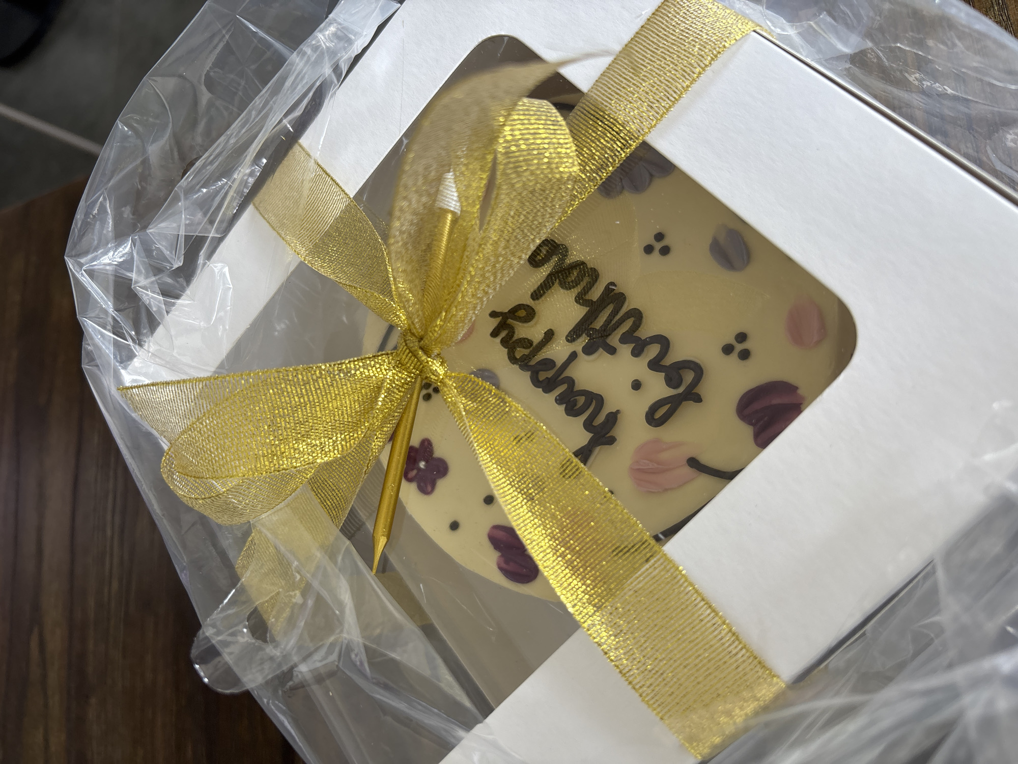 The image shows a white box, likely containing a cake or dessert, wrapped in clear plastic and tied with a gold ribbon. The box has a window revealing a decorated cake with "Happy Birthday" written in black frosting. There are also floral decorations on the cake. A gold-colored candle is inserted into the cake. The box is sitting on a dark wooden surface. The photograph appears to be taken indoors. No specific location within Kuta Selatan, Indonesia is discernable from the image. No people are visible in the image.