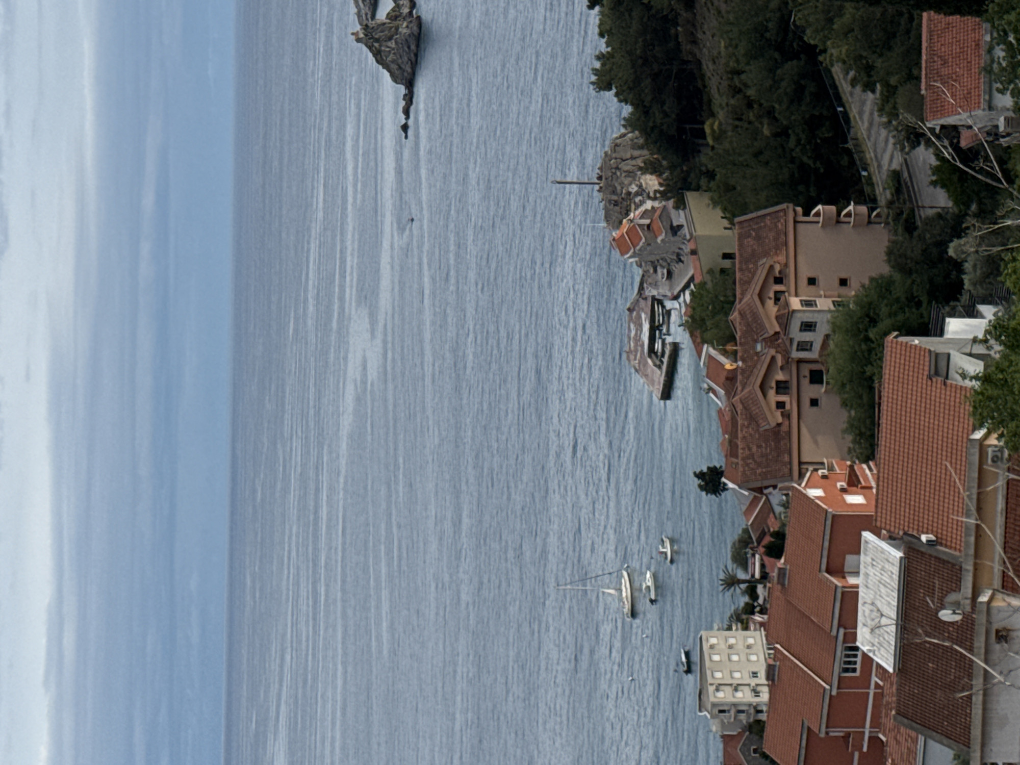 An aerial, rotated view of a coastal city on the Adriatic Sea. In the foreground, there are buildings with red-tiled roofs, trees, and a few communication towers. The architecture style appears to be typical Mediterranean. The sea occupies a large portion of the view with several sailboats visible. Along the coast, there are more buildings and trees. In the far distance, there is a rocky cliff near the horizon. The sky is partially covered with clouds.