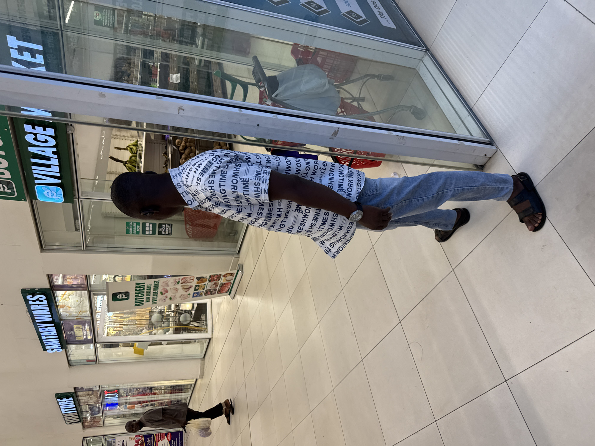 The image depicts the interior of a shopping mall in Abuja, Nigeria, evidenced by the "Village Market" sign. A man wearing jeans and a shirt with text prints stands in the mall's tiled walkway. Storefronts displaying items such as sanitary wares, furniture, and butchery products are visible on the left. Another person can be seen walking with a shopping bag at the end of the hallway.