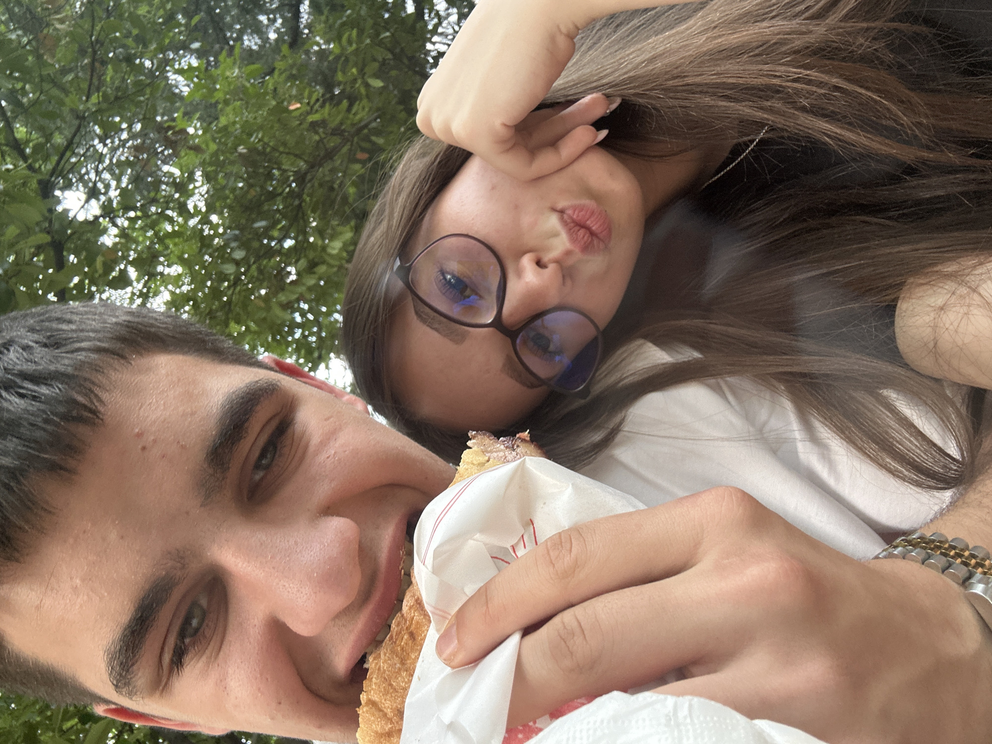 This is a casual, low-angle selfie-style photograph capturing two young individuals outdoors, likely in a park or a green area in Blagoevgrad, Bulgaria. The shot is taken from a perspective looking upwards, with the background consisting of lush green tree foliage and glimpses of a bright sky.
In the foreground, a young man with short dark hair and green eyes is visible, smiling slightly. He is in the process of taking a bite from a pastry or sandwich, which is partially wrapped in white paper adorned with thin red stripes. His left hand holds the food, while his right wrist, partially visible, sports a two-tone metallic watch with gold and silver-colored links.
Behind and slightly above him, a young woman with long, straight brown hair is present. She is wearing dark-rimmed glasses, which show reflections of blue light, and is looking towards the camera with a "duck face" or a puckered-lip expression. Her right hand is raised, partially covering her head, and a subtle silver chain earring can be seen near her ear. She appears to be wearing a white top.
The overall scene suggests a relaxed, playful moment shared between the two, possibly during a daytime outing or picnic. The light appears natural, indicating it is daytime, and the expressions convey a casual and affectionate mood. The food wrapping has red striped markings but no clearly legible text.