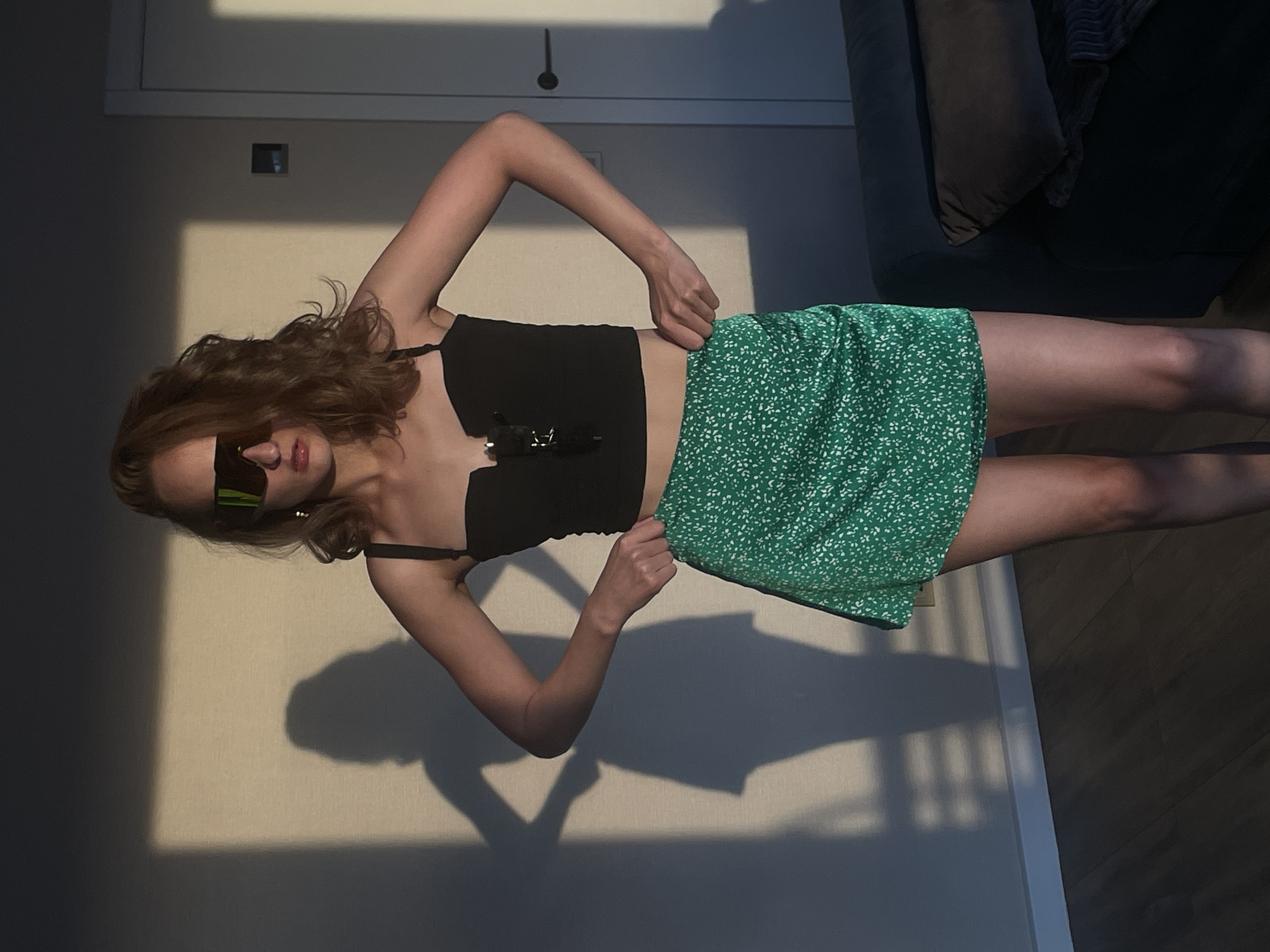 A woman wearing sunglasses, a black top, and a green floral skirt is standing indoors with one hand on her skirt. She is positioned in front of a light-colored wall with shadows cast upon it, and next to a dark-colored couch. There is no indication that this image was taken in Подшивалово, Russia.