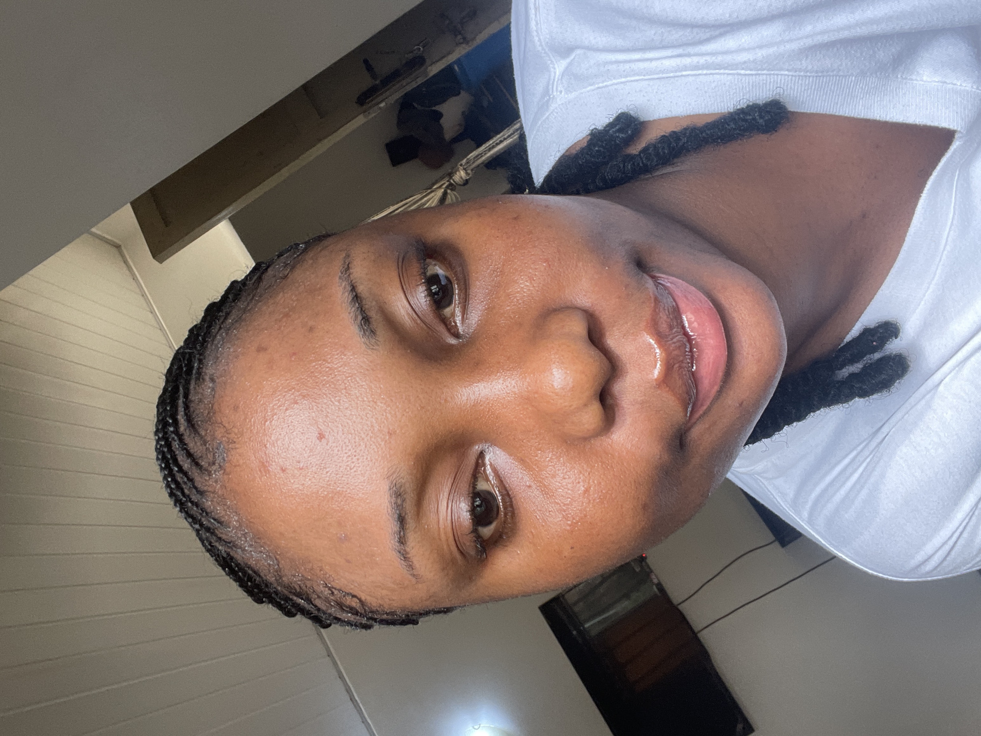 A close-up, rotated image depicts a woman of color with dark, braided hair. She is wearing a white shirt. The background shows a room with light-colored walls and a wooden beam structure near the ceiling. The location, Abuja, Nigeria, is not verifiable from this image alone.