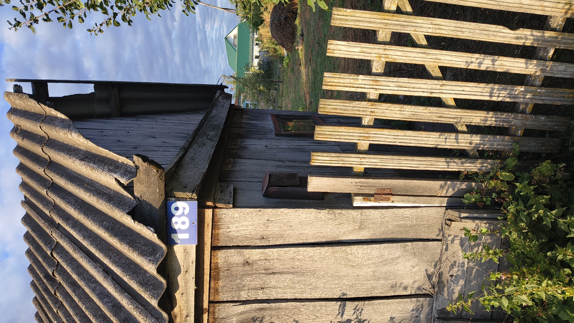 The image shows a weathered wooden outbuilding with a corrugated roof in Zemlyanichnyi, Russia. A blue sign with the number "189" is affixed to the front of the building. A rustic wooden fence runs along the foreground, with green foliage growing around its base. In the background, a house with a green roof is visible, along with a haystack. The scene is bathed in the warm light of either sunrise or sunset.