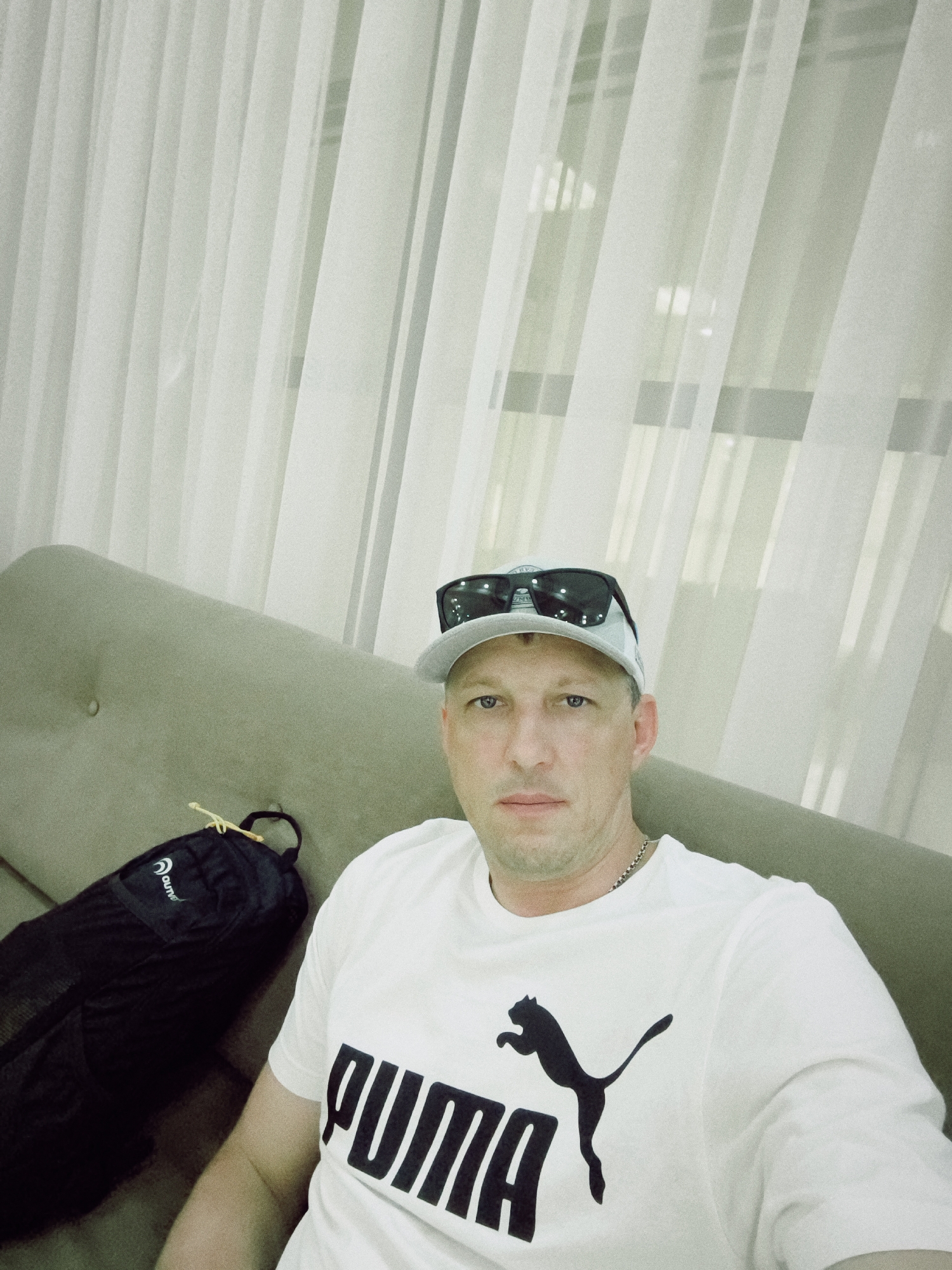 A man is sitting on a sofa in front of a window with white curtains. He is wearing a white t-shirt with the Puma logo on it, a baseball cap, and sunglasses. A black backpack is on the sofa next to him. The setting appears to be an indoor space, possibly a hotel room or a lounge area. There are no clear indicators in the image to pinpoint the specific location within Sochi, Russia.