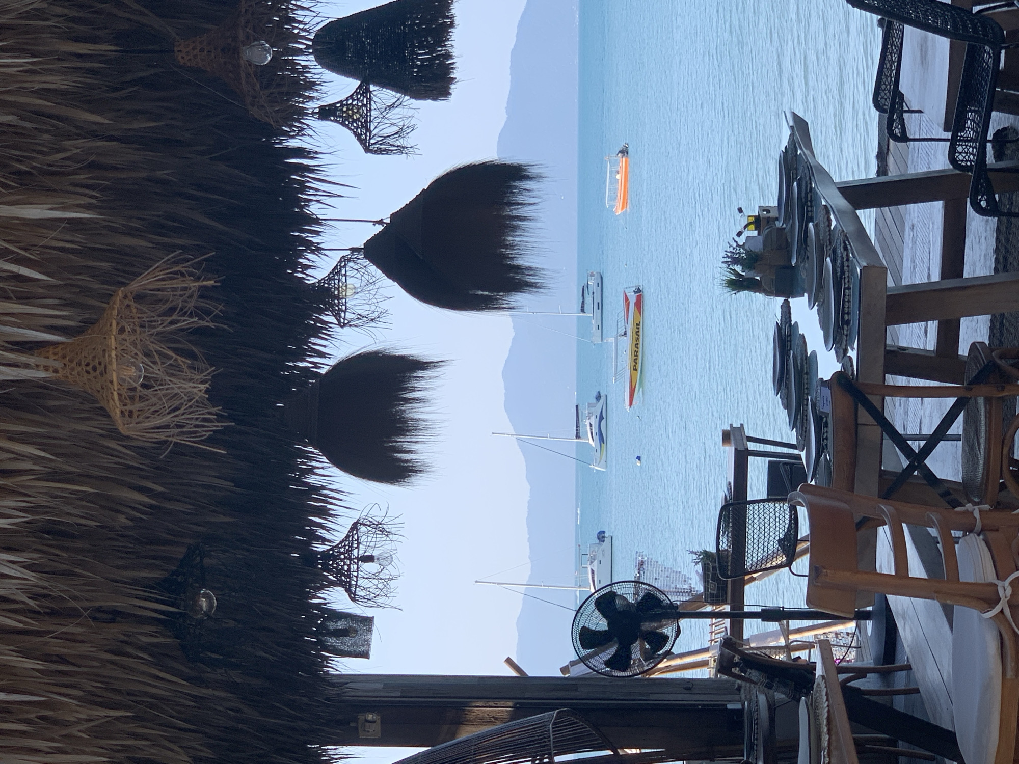The image shows a view from a thatched-roof outdoor dining area in Ko Samui District, Thailand, overlooking the ocean. In the foreground, several wooden tables and chairs are set for dining, with plates and placemats arranged. A standing fan is visible on the left. Hanging from the thatched ceiling are various woven light fixtures. Beyond the dining area, the turquoise ocean stretches out, dotted with sailboats and smaller boats. In the distance, a hazy mountain range is visible against a clear sky. The scene suggests a relaxed, tropical restaurant setting by the sea.