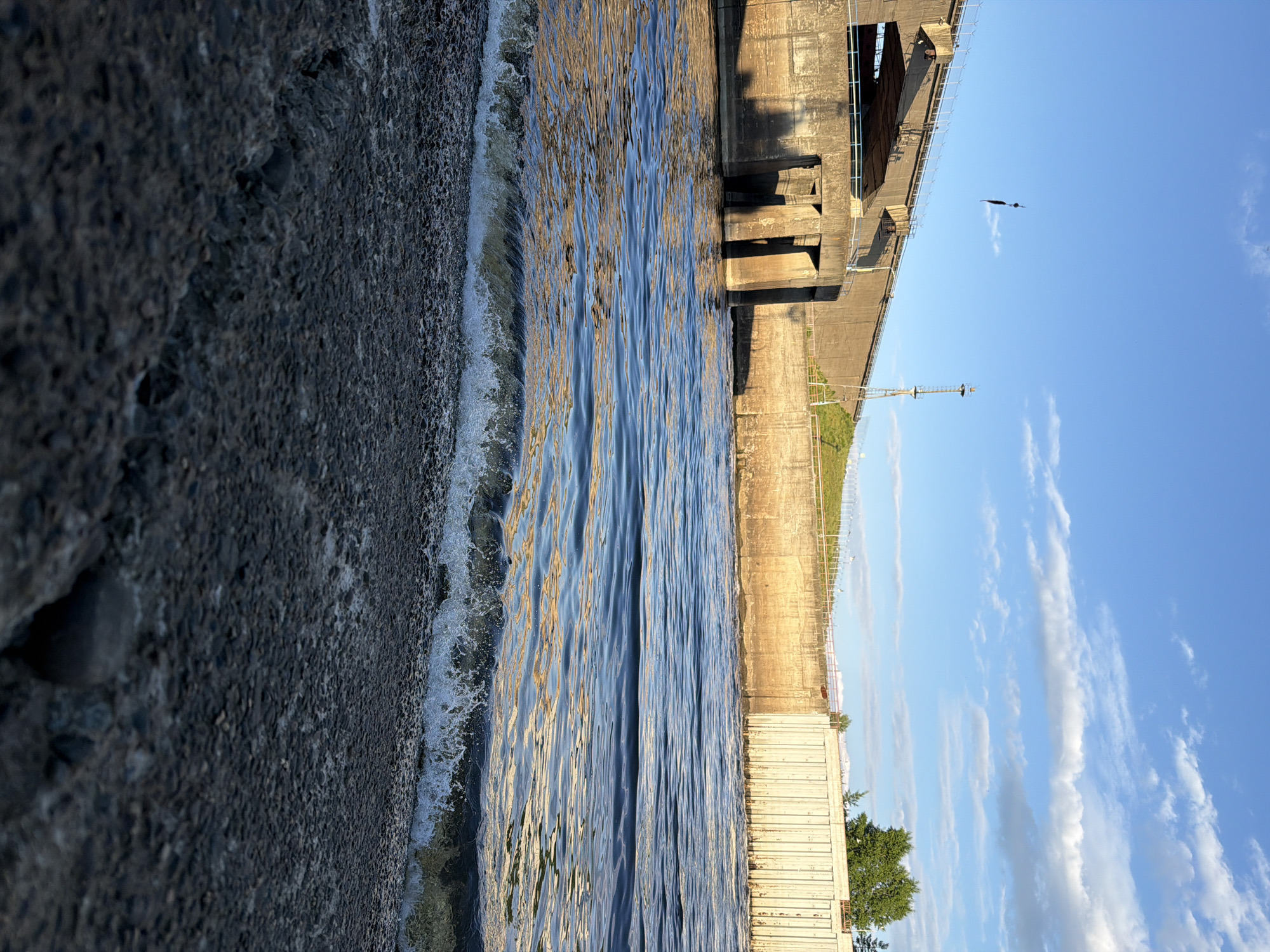 The image depicts a body of water flowing towards a pebbled shore in front of a concrete dam structure. The dam features several concrete pillars and a metal gate system on the right side. In the background, there's a large concrete wall and a sloped grassy area with a fence. The sky is blue with scattered white clouds. A dark object, possibly a piece of equipment, is suspended from a cable above the dam. The scene appears to be located at the Votkinsk Hydroelectric Power Station dam on the Kama River, which serves the city of Chaikovsky, Russia. The water is rippling, with a small wave breaking at the shore. The lighting suggests it is late afternoon or early evening, with the sun casting shadows on the dam.