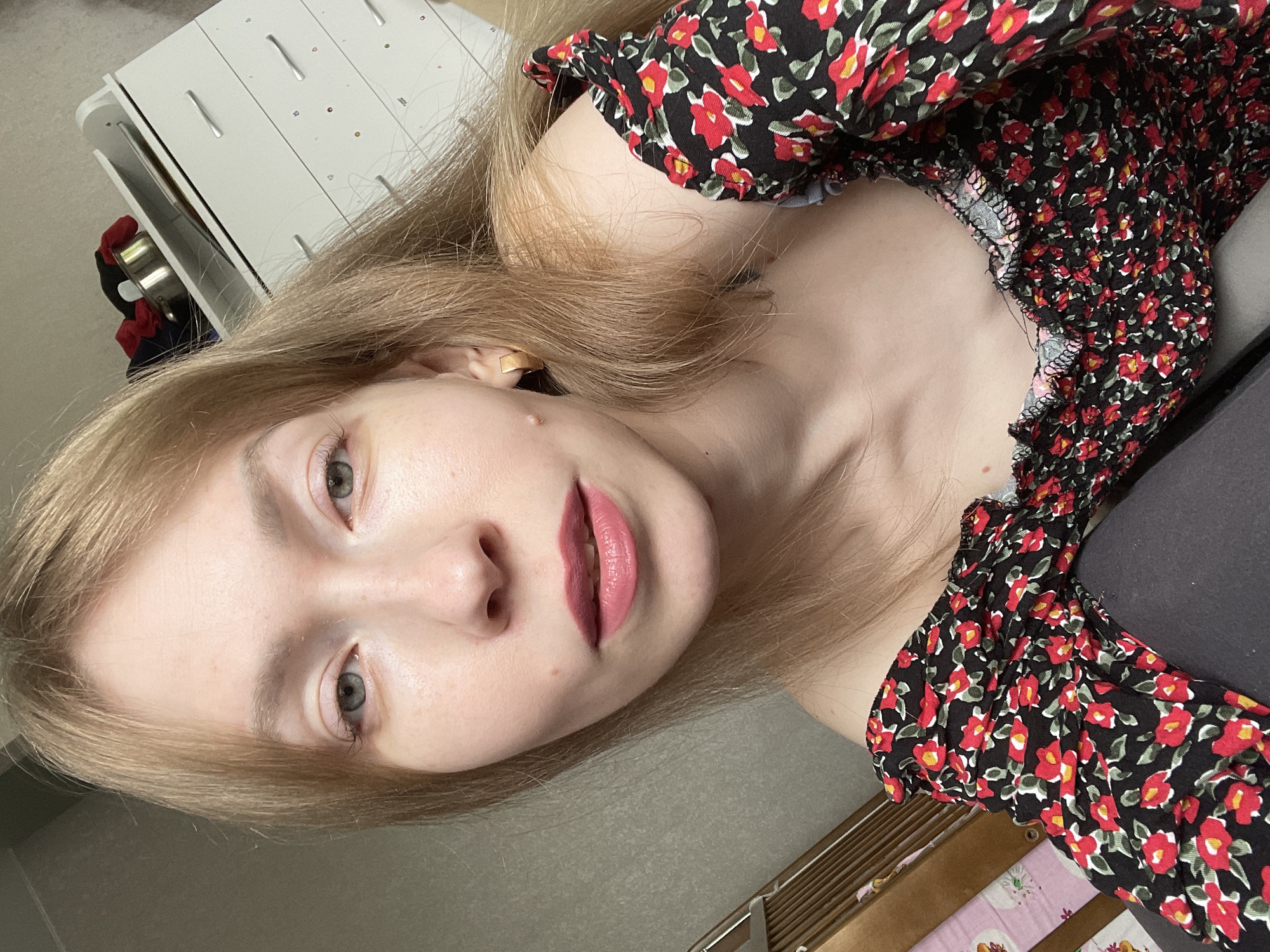 The image shows a woman with light blonde hair, fair skin, and gray eyes looking directly at the camera. She has a visible mole on her cheek and is wearing lipstick. She is wearing a black floral dress with small red flowers. Behind her, there is a white cabinet with metal handles and a gray wall. There is also a partially visible drawer unit with pink and white patterned fabric. There is no information to suggest the location is in Perm, Russia.