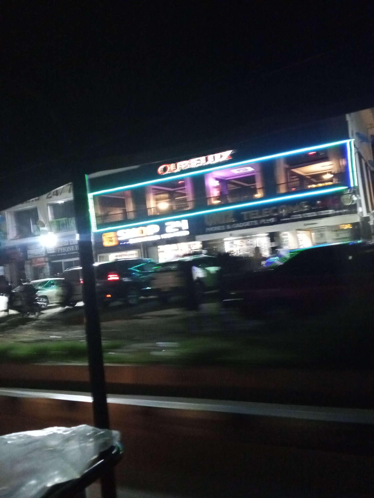 The image, taken at night in Lokoja, Nigeria, shows a brightly lit commercial building. The building has multiple stories, with neon blue and purple lights outlining its facade. A prominent sign reads "OURELUX" in large letters, with another sign below it saying "SHOP 21". Additional signs for "OMIZ TELECOM" and "PHONES & GADGETS PLUS" are also visible, suggesting the building houses various businesses. Several cars are parked or driving in front of the building, their headlights and taillights illuminated. The overall scene is characteristic of a bustling urban commercial district at night.