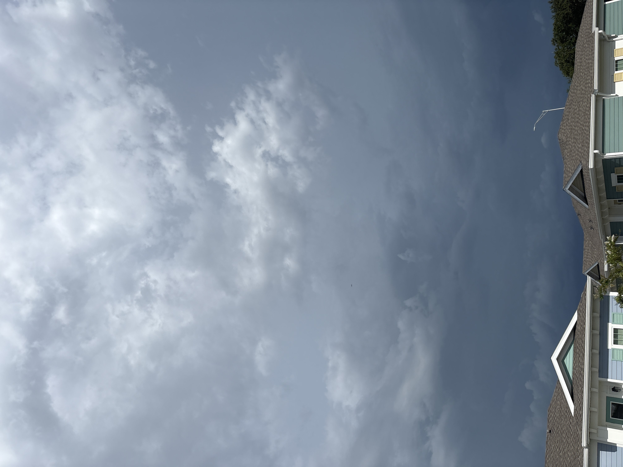 An overcast sky looms above two houses in Sarasota, Florida. The clouds are dark and heavy, suggesting an impending storm, with lighter clouds scattered across the upper portion of the frame. The houses, painted in light blue and white with light teal accents, have gray shingled roofs. A streetlight is visible above the right house. The image is captured from a low angle, emphasizing the dramatic sky. There are no people or animals visible in the image.