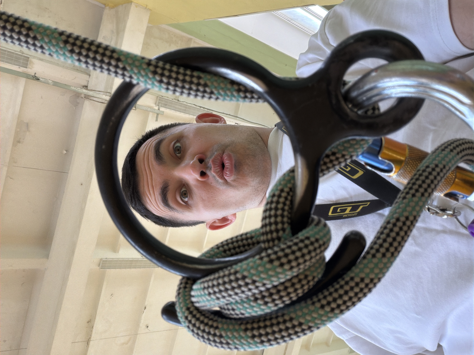 A man, face contorted in a pucker, is the primary subject in a low-angle, close-up shot. He's framed by climbing equipment, including a black metal device, a silver carabiner, and a thick rope looped through the equipment. A "GT Bicycles" lanyard or strap is also visible. The background is out of focus but appears to be an interior space with ceiling support structures painted in light colors. The photograph appears to have been taken indoors, possibly in a training or climbing facility in Izhevsk, Russia.
