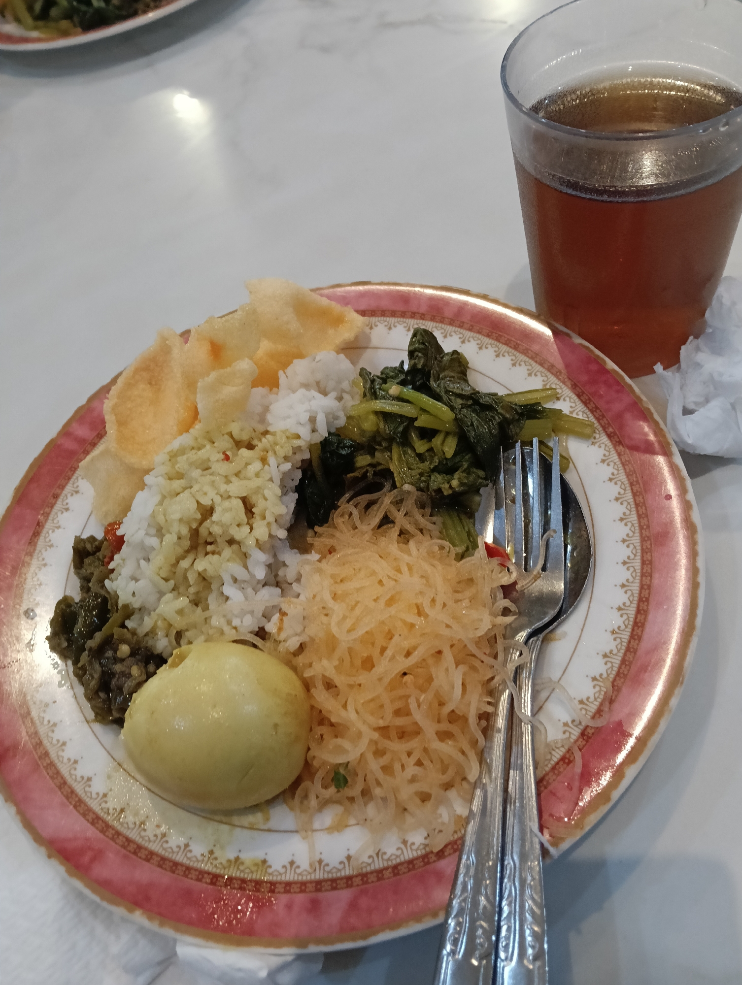 An overhead shot shows a meal served on a decorative plate next to a glass of iced tea. The plate is filled with white rice, a hard-boiled egg, a serving of greens, fried noodles, and several pieces of crackers. A fork and spoon are placed on the right side of the plate, partially submerged in the noodles. The setting appears to be a casual dining establishment, possibly in Cikawung, Indonesia, given the context provided. The table surface is a pale, reflective material, suggesting it might be marble or a similar stone. A crumpled white napkin is visible to the right of the glass of tea. The lighting is bright, casting subtle shadows and highlighting the textures of the food and plate.