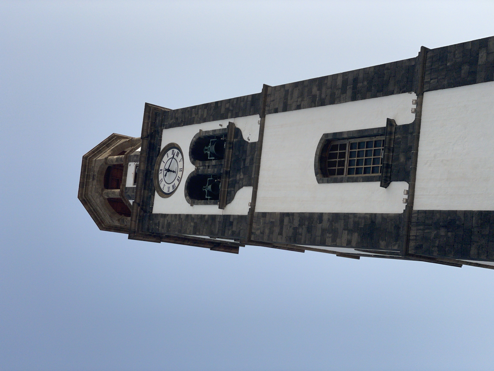 The image shows a low-angle view of a church tower against a clear sky. The tower has a white facade with dark stone trim. It features a clock face, two bell openings with bells visible inside, and a window with a grid pattern. The tower appears to be located in the city of Santa Cruz de Tenerife, Spain, based on architectural style. No people are visible.