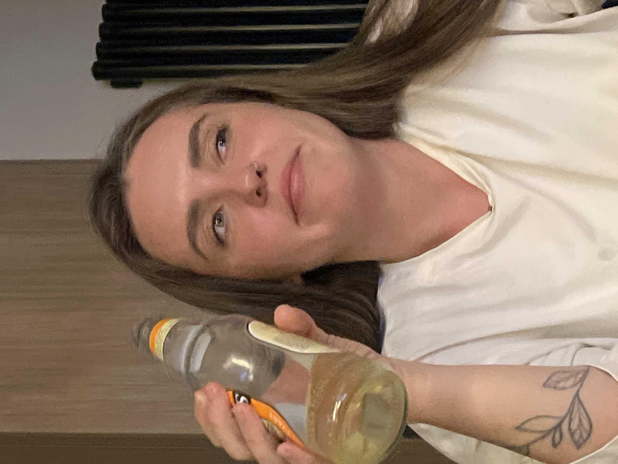 A young woman is reclining, holding a bottle of liquid. She has light skin, brown hair, and a nose ring. She wears a white garment, and a floral tattoo is visible on her right forearm. The bottle contains a yellow liquid, and the label is orange. The background is a mix of light and dark surfaces, including what appears to be a wooden surface and a dark, possibly metal, structure. There is no indication of location in the city of Рубцово, Russia.
