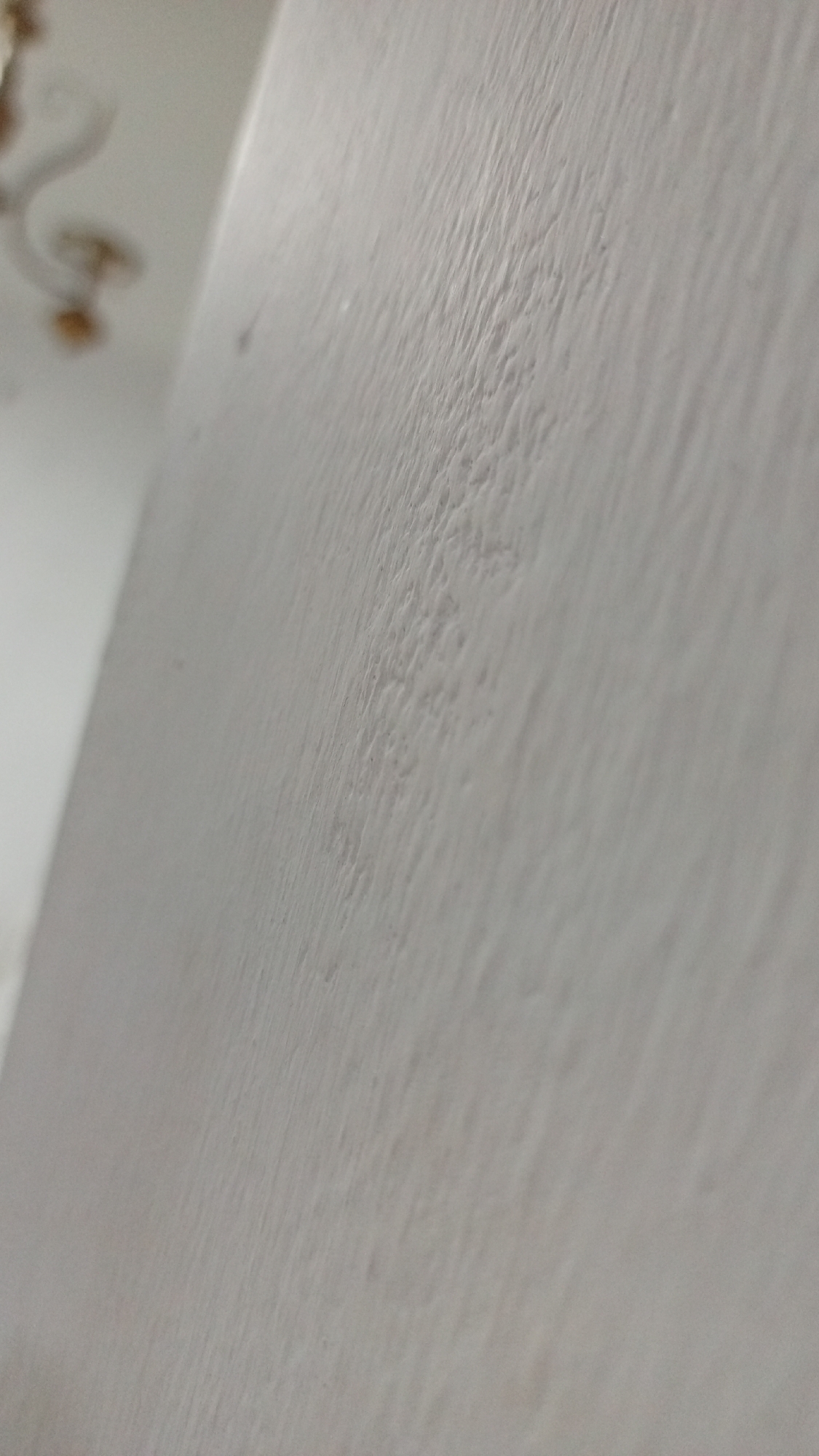 The image shows a close-up of a textured, off-white wall. In the upper left corner, a blurred portion of a ceiling with decorative elements, likely from a light fixture, is visible. The texture of the wall consists of a pattern of ridges and valleys. The image provides insufficient information to determine the specific location in Los Robles, Venezuela, or to identify any people, subjects, objects, actions, interactions, or specific environmental details beyond the wall and ceiling.