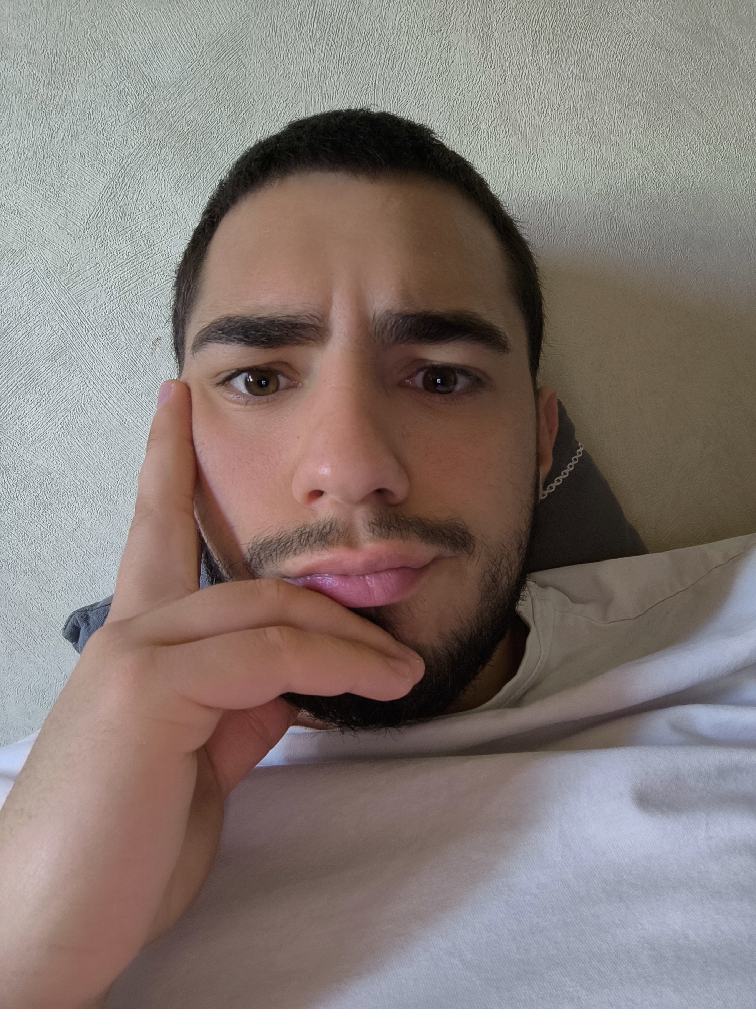 A selfie-style photograph captures a man in a close-up shot, likely taken indoors in Schiltigheim, France. The man has short, dark hair, dark eyebrows, and brown eyes, looking directly at the camera. He sports light facial hair, a tan complexion, and pinkish lips. His left hand is raised, with his index finger resting against his temple and cheek, and his thumb positioned near his chin, creating a pensive or contemplative expression. He is wearing a plain white t-shirt. A notable detail is a silver chain earring visible on his right ear, contrasting with the dark grey pillow or cushion behind him. The background features a light-colored, textured wall with a subtle patterned design, consistent across the various crops. The crops provide closer views of his facial features, the texture of the wall, and the earring. There is no visible text in the image. The overall impression is a casual, indoor setting.