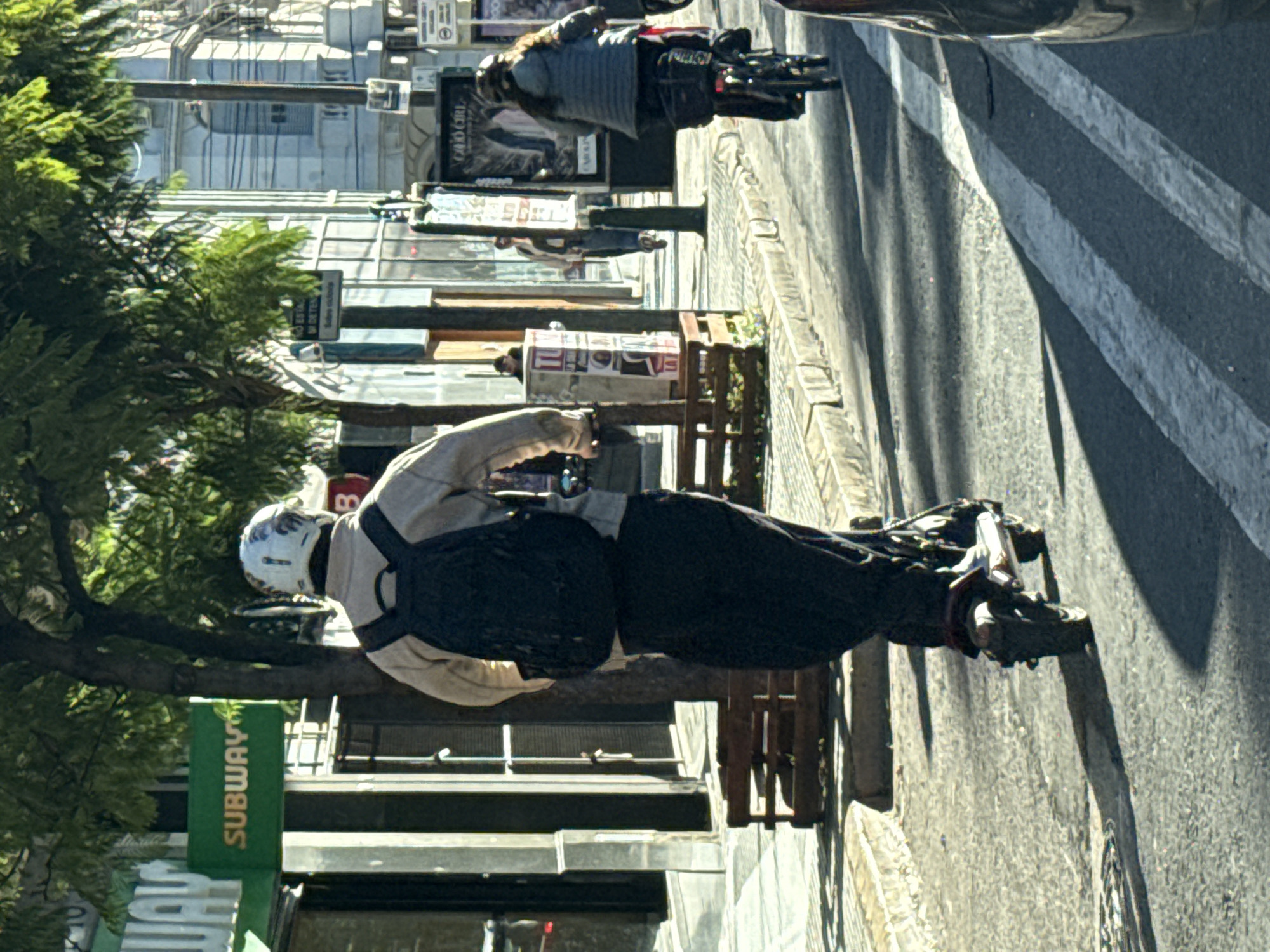 This street-level photograph, taken in Buenos Aires, Argentina, captures a bright, sunny urban scene. In the foreground, a person is standing next to an electric scooter or similar personal mobility device. They wear a white helmet with a design, a light-colored jacket, dark pants, and a dark backpack. Their posture suggests they are paused, possibly looking down at the device or their phone. The scooter has visible suspension and foot platforms.
Further down the sun-drenched street, another individual is seen on a similar electric scooter, wearing a dark jacket, moving away from the viewer. The setting is a bustling city sidewalk, lined with various commercial buildings, some with large glass windows reflecting the bright sky. Trees with green foliage cast shadows, and wooden benches are visible along the sidewalk.
Strong sunlight creates sharp, distinct shadows across the asphalt road, which features a white crosswalk marking. The time of day appears to be midday or early afternoon, given the intensity of the light.
Visible text includes a prominent green "SUBWAY" sign, indicating a fast-food restaurant. Another sign in the background, difficult to fully decipher, appears to say "CADOURE". A white rectangular street sign also displays Spanish text, partially readable as "NO ESTAC... W DE ESQ...", likely an instruction regarding parking restrictions near a corner. The overall impression is one of modern city life, with personal electric transport being a common sight.