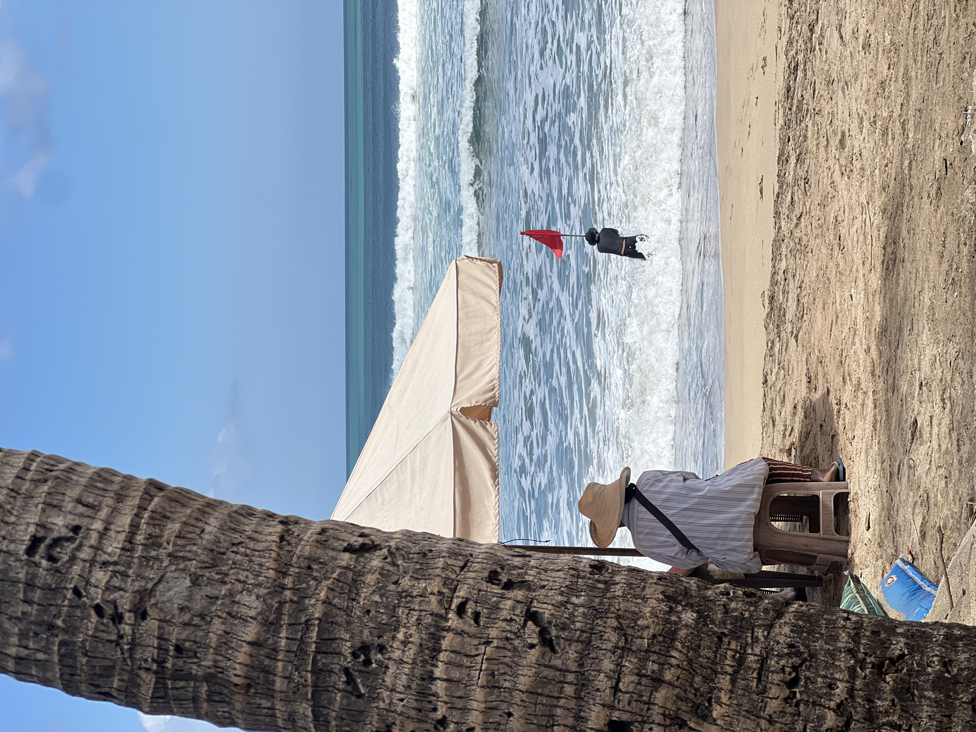 A person wearing a wide-brimmed hat and a striped shirt sits on a stool under a beach umbrella on the sandy shore of Kuta Beach, Indonesia. The person faces the ocean, where waves crash onto the beach. In the water, a person in dark clothing holds a red flag aloft. The scene is framed by the rough bark of a palm tree trunk on the left side of the image. The sky is a clear blue with a few wispy clouds.