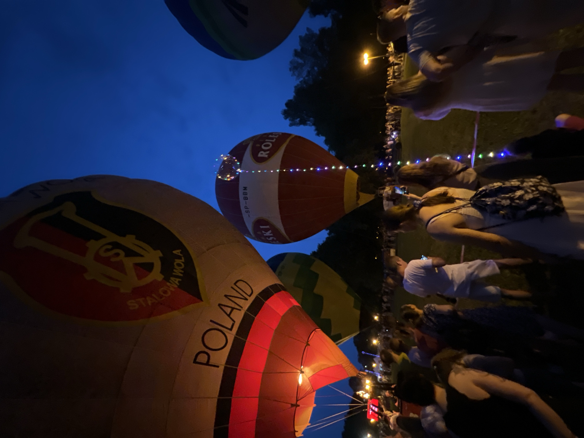 This image depicts a nighttime hot air balloon event in Tychy, Poland. In the foreground, a large hot air balloon with the word "POLAND" printed on it is prominently displayed, illuminated by its internal lights. To the left, another balloon features a crest with the text "STALOWA WOLA" and a stylized symbol. In the center, a red and white balloon is adorned with multicolored lights cascading down its side, and the word "ROLEX" is visible. In the background, a crowd of people is gathered, silhouetted against the twilight sky and illuminated by distant lights and the glow of the balloons. Trees form a dark backdrop against the deep blue sky, and streetlights are visible in the distance. The overall scene captures a festive atmosphere during a twilight hot air balloon festival.