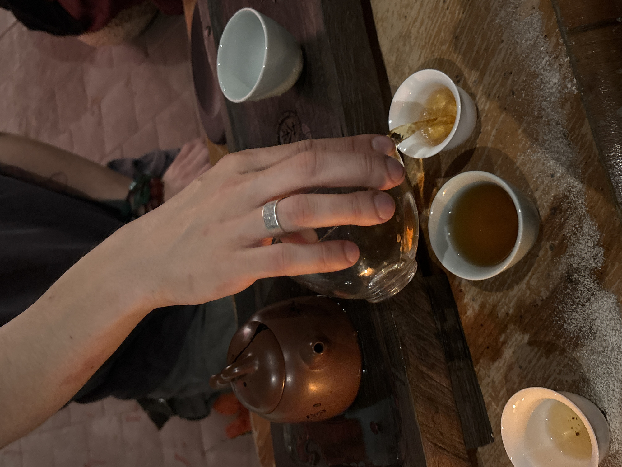 A person is pouring tea from a glass pitcher into a white teacup on a wooden surface. A brown teapot is nearby. Three other white teacups are present, two containing tea and one empty. The wooden surface has white particles scattered on it. The person wears a silver ring on their left ring finger and a green bracelet on their left wrist. The location is potentially Saint Petersburg, Russia, based on the image composition and subject matter of tea pouring/drinking.