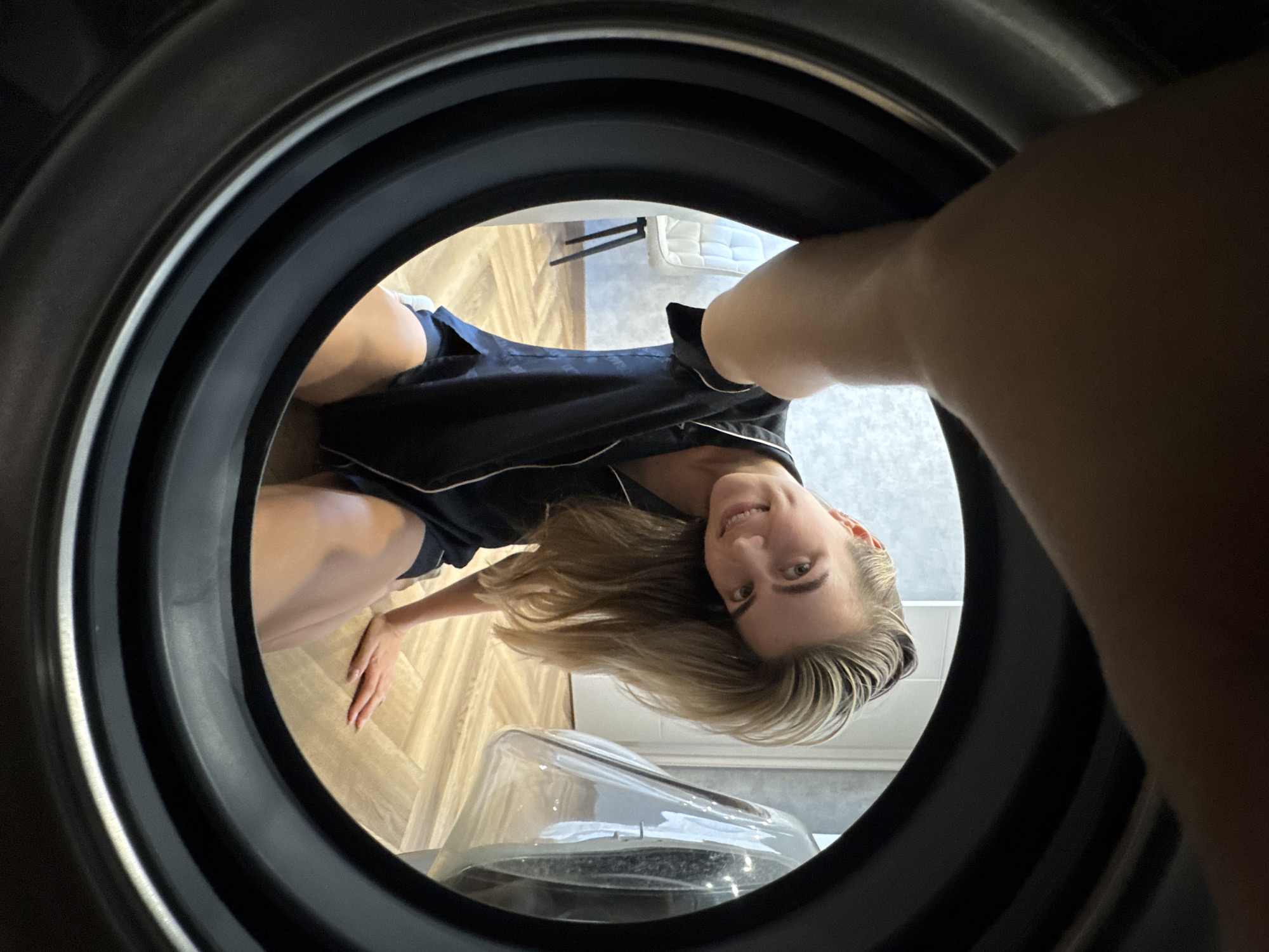 A person is viewed from inside a dark, circular opening, possibly a washing machine or dryer drum. The person, appearing female with fair skin and light blonde hair, is wearing a dark blue pajama set with white piping. She is smiling and looking directly at the camera. The interior background consists of a light-colored, wood-patterned floor, a clear object that appears to be a humidifier, and part of a white cushion or mattress with a black metal frame. The location appears to be indoors, possibly a residence in the city of Unknown, Unknown.