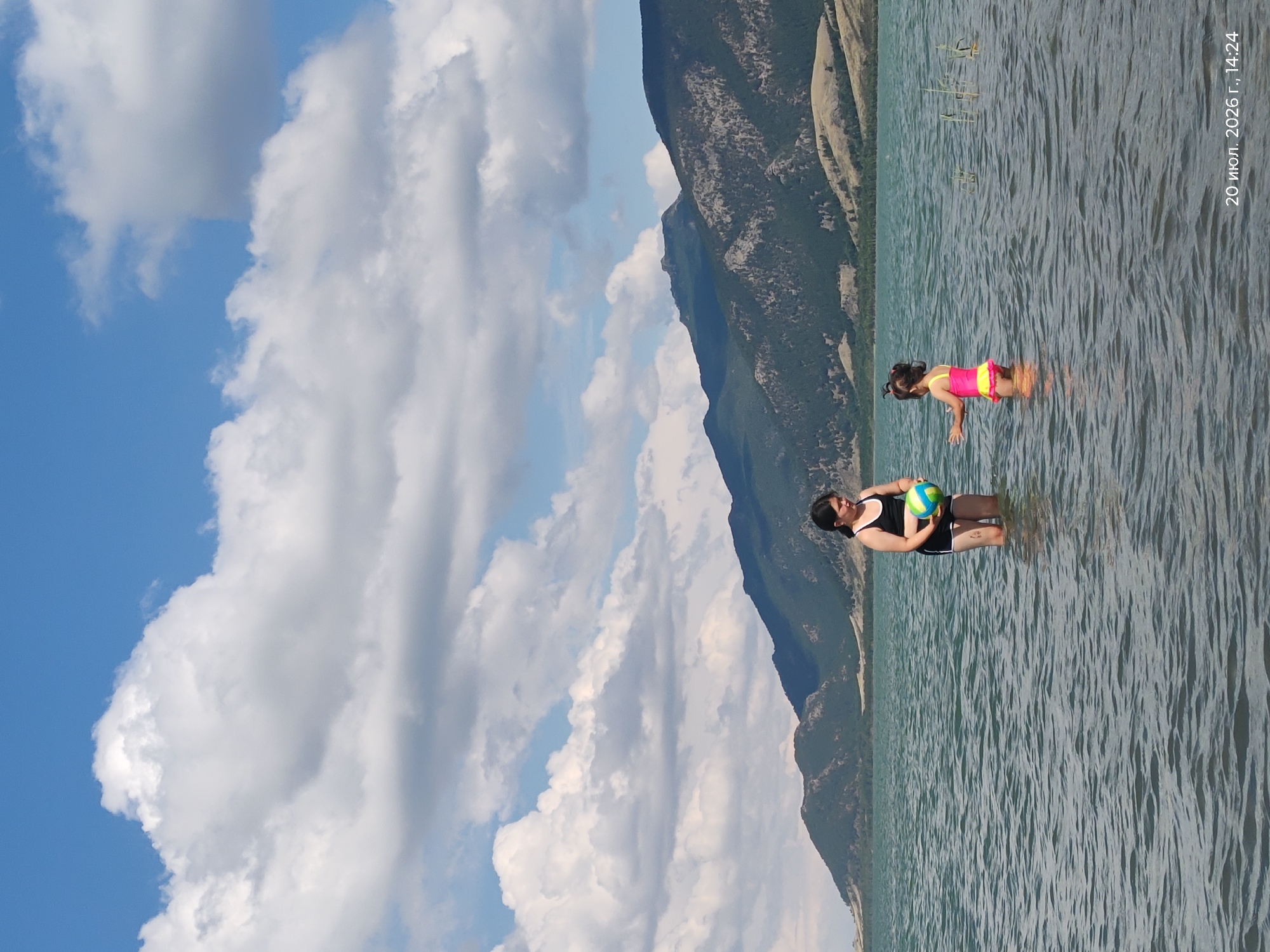 Two children are standing in the water of a large lake, with mountains in the background. The child on the left is older, wearing a black swimsuit and holding a multi-colored ball. The child on the right is younger, wearing a pink and yellow swimsuit. The water is calm with small ripples. The sky is blue with fluffy white clouds. The date stamp in the lower right corner reads "20 июл. 2026 г., 14:24". The location is the city of Akzhol, Kazakhstan.