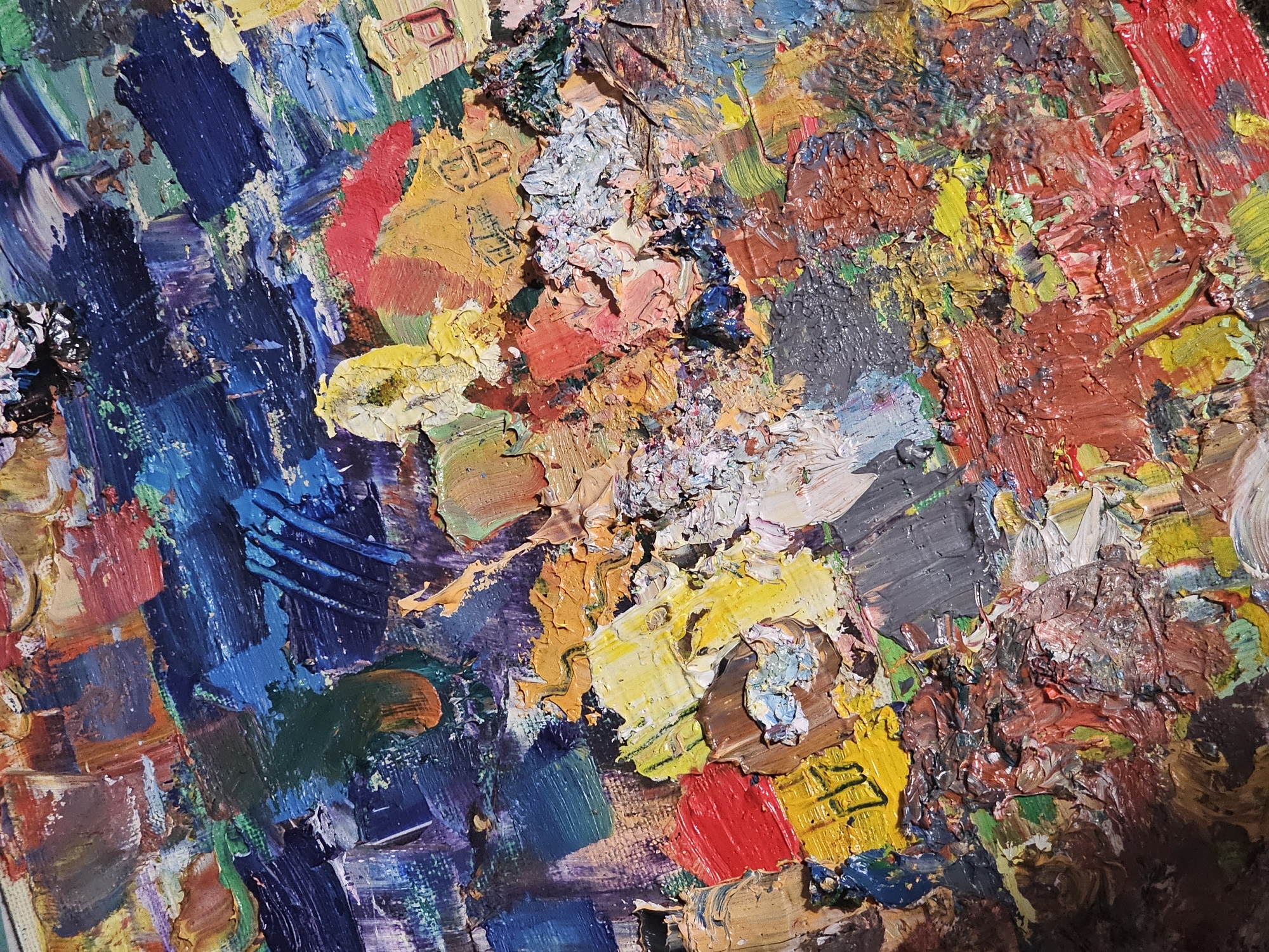The image is a close-up of an abstract painting, likely oil or acrylic, characterized by thick, textured brushstrokes. Dominant colors include blue, yellow, red, brown, and gray, blended and layered in a chaotic arrangement. There are no discernible figures or recognizable objects. The lighting appears to be from above, casting shadows that accentuate the texture. The image provides no context related to the city of Plovdiv, Bulgaria.