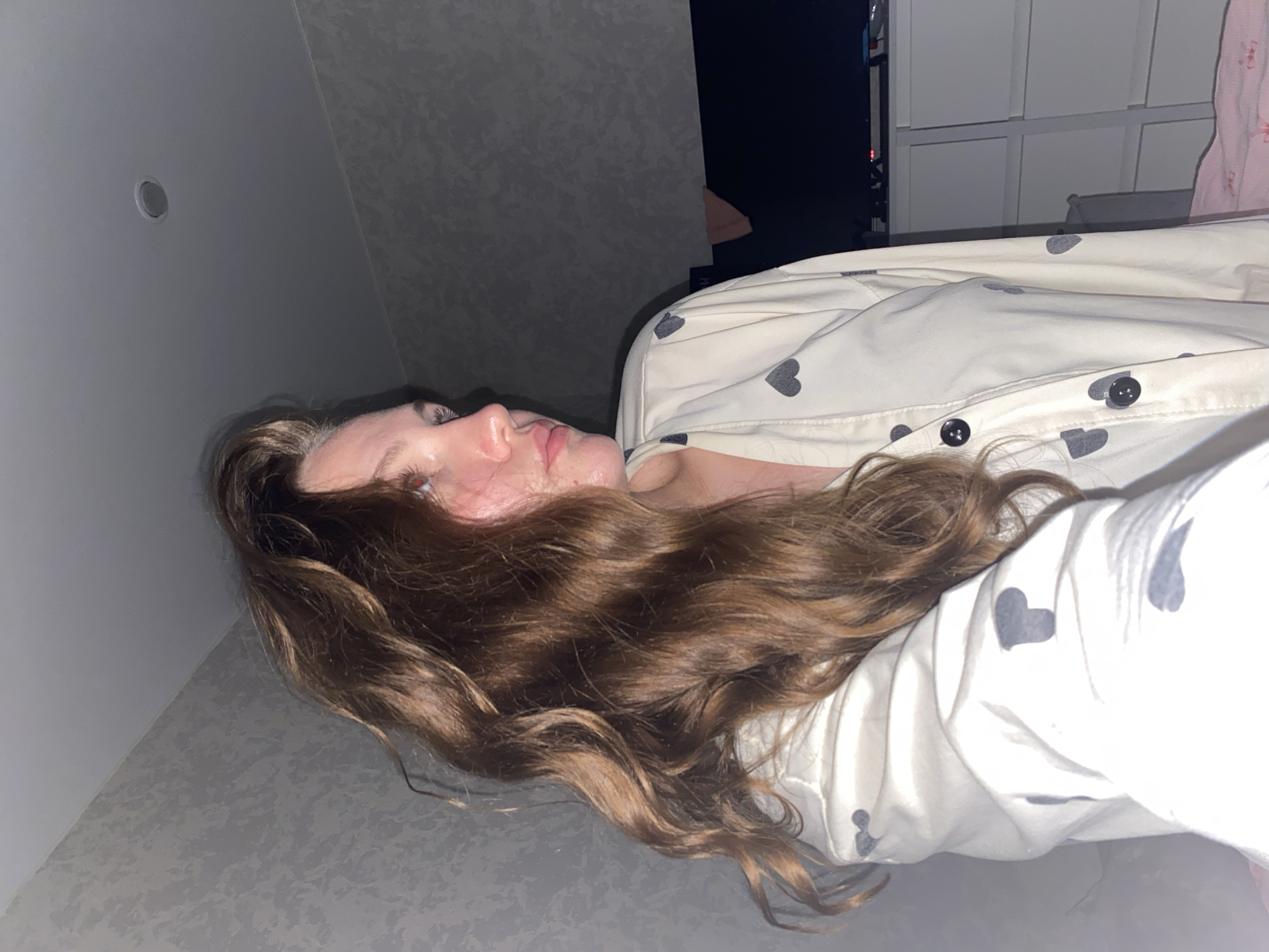 A woman is lying on a gray surface. She has long, brown, wavy hair. She is wearing a light-colored button-down shirt with gray heart prints. Behind her is a light gray wall with a recessed light, as well as a dark space and light-colored furniture. The location is unknown.