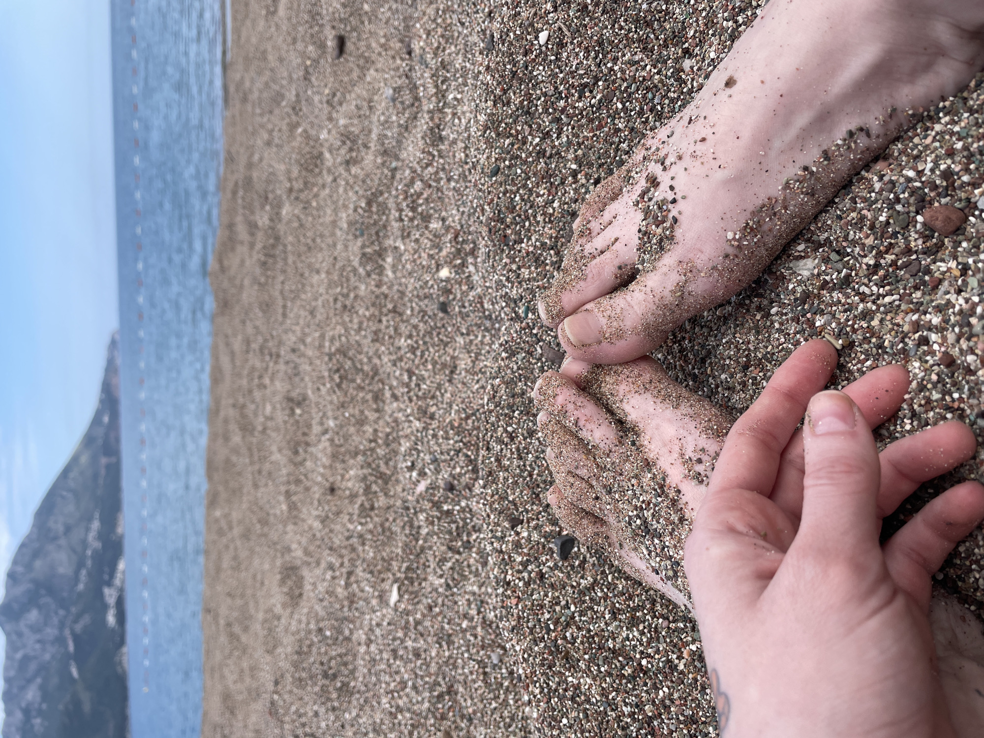 A ground-level, outdoor shot features a pair of feet and a hand resting on a coarse, grainy beach, with the sea and distant cliffs visible in the background. The feet and hand are partially covered in sand. The location is Bečići, Montenegro. The composition suggests the subject is at rest, interacting with the beach environment.