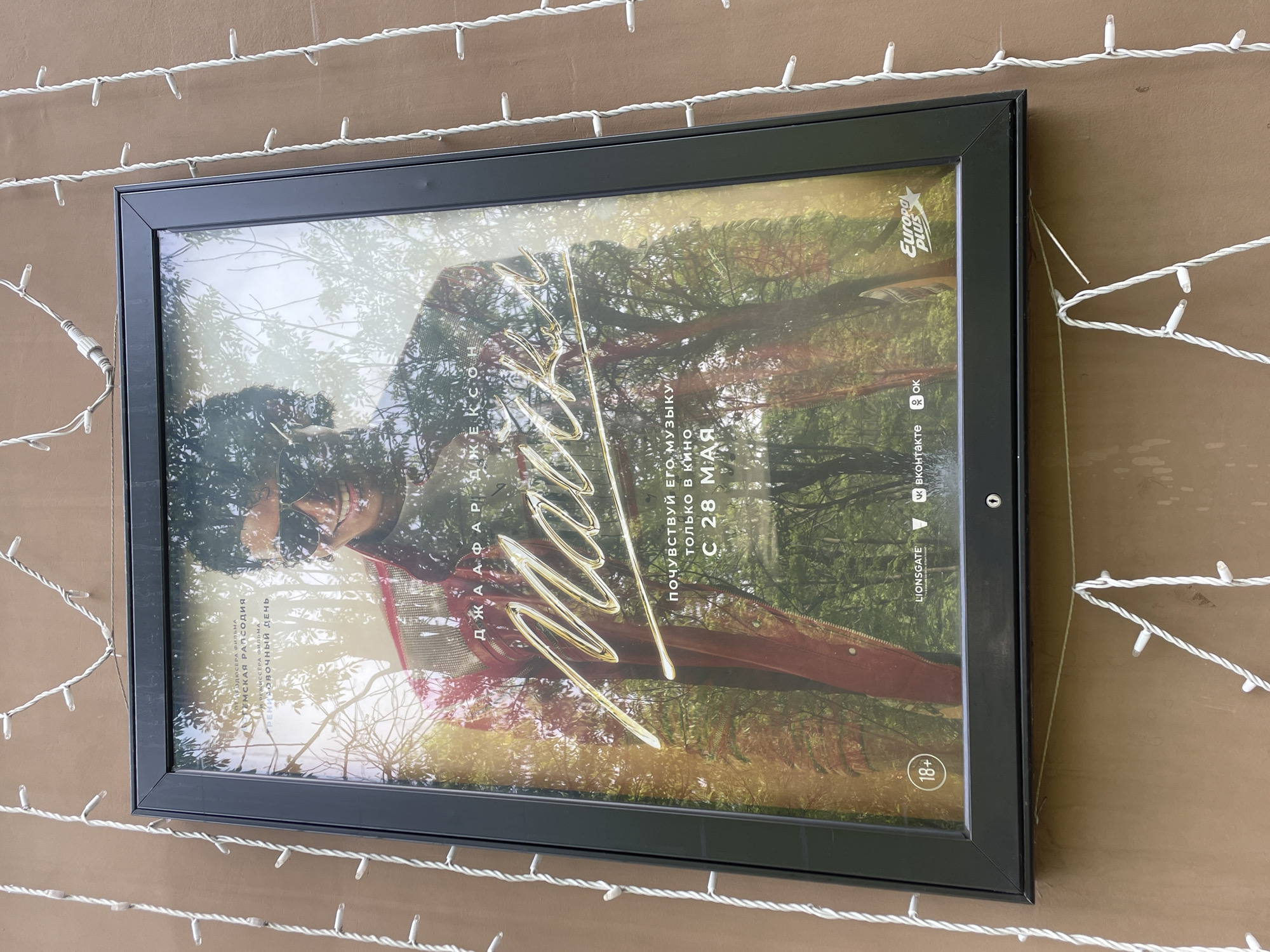 The image shows a framed poster for the movie "Michael Jackson" hanging on a brown wall decorated with strings of white lights. The poster features a blurred image of Michael Jackson with a forest backdrop, overlaid with a golden signature. Text on the poster is in Russian, including movie production details and the release date of May 28th. Logos for Europa Plus, Lionsgate, and social media platforms VKontakte and OK are also visible. The frame is black, and a small security lock is attached to the right side. The scene is in Khimki, Russia.