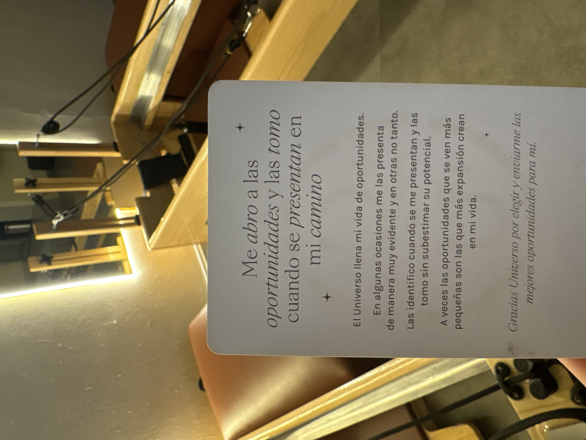 The image shows a card with text on it, held up in front of what appears to be a Pilates studio. The card has a light gray background with rounded corners. The text on the card is in Spanish and is in a decorative font. The top of the card has a title that reads "Me abro a las oportunidades y las tomo cuando se presentan en mi camino." Below this, there are several paragraphs of text. The background of the image shows workout equipment, likely Reformers, in a bright, modern space with a large illuminated mirror. The lighting suggests an indoor setting, possibly a gym or fitness studio. There is no indication of the location being Mexico City, Mexico.