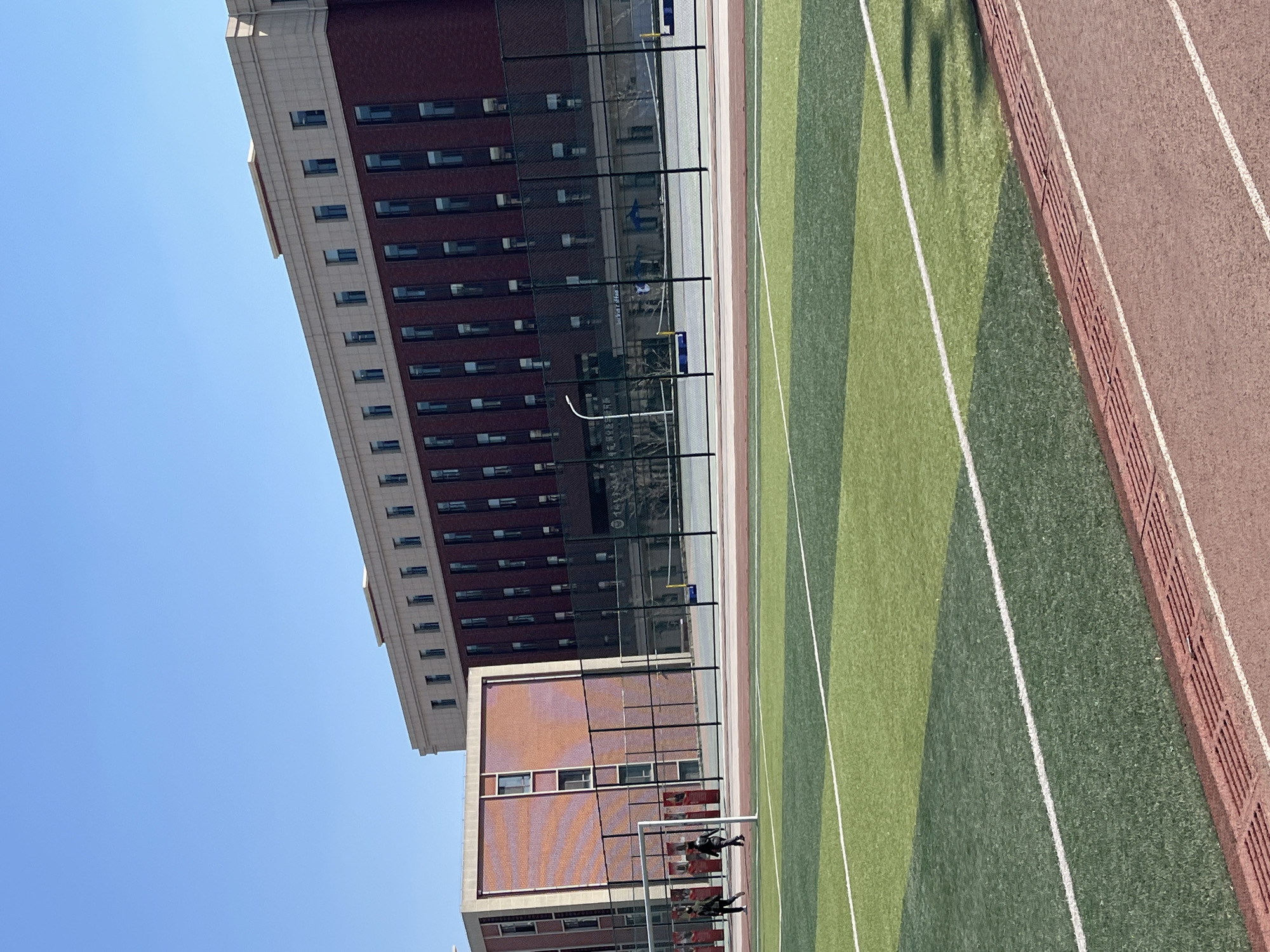The image shows an outdoor scene in Changchun City, China. The view is angled upwards, revealing a blue sky at the top left. Two buildings are visible. The taller building is a multi-story structure with a red brick facade, light-colored trim, and numerous rectangular windows. The shorter building is a brick structure. Between the buildings and the viewer is a sports field with alternating stripes of light and dark green turf, separated by white lines. A red running track borders the field. A black chain-link fence runs along the edge of the field. Figures, appearing to be people, are visible behind a soccer goal.